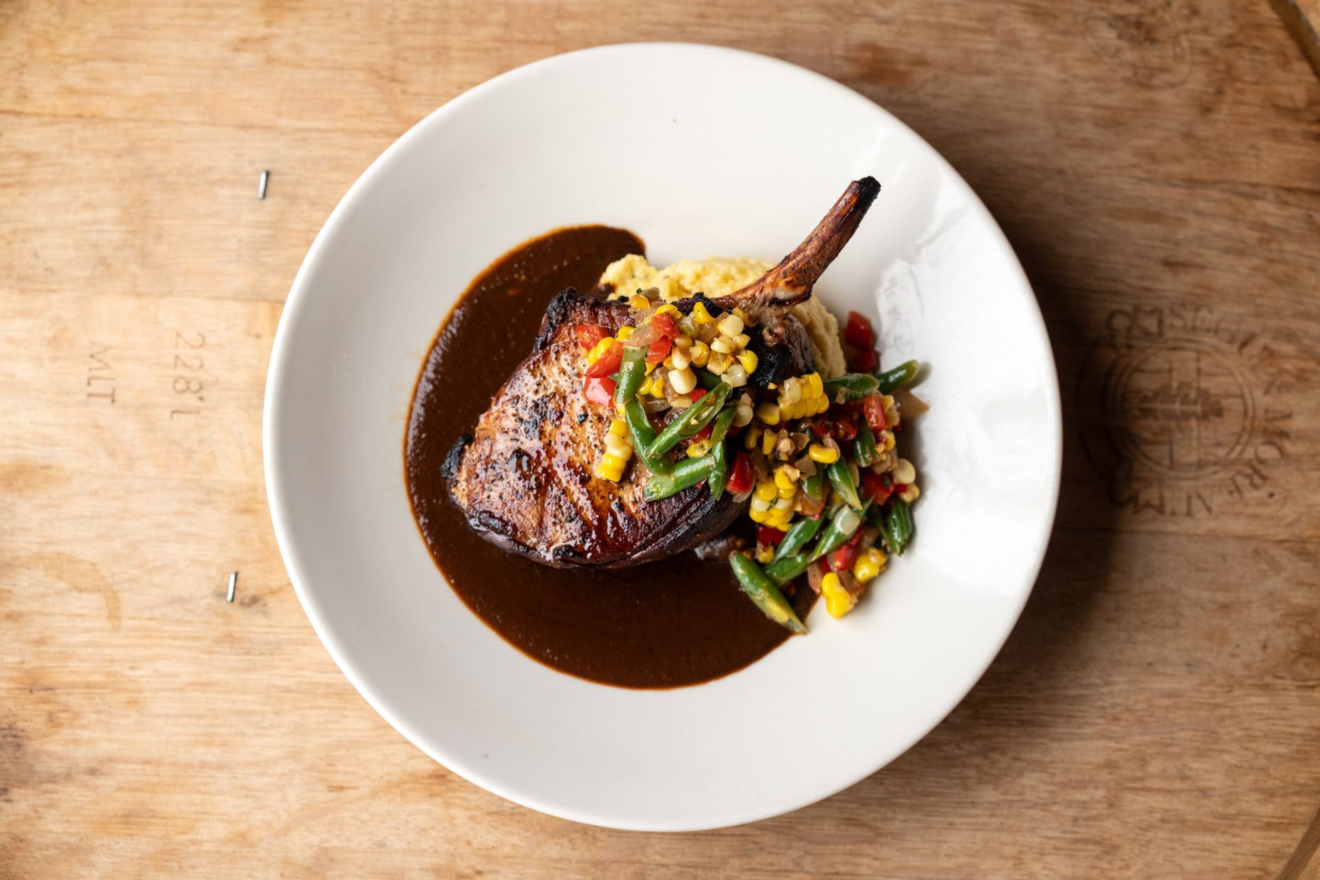 Pork chop with sauce and a vegetable medley on a white plate, set on a wooden surface.