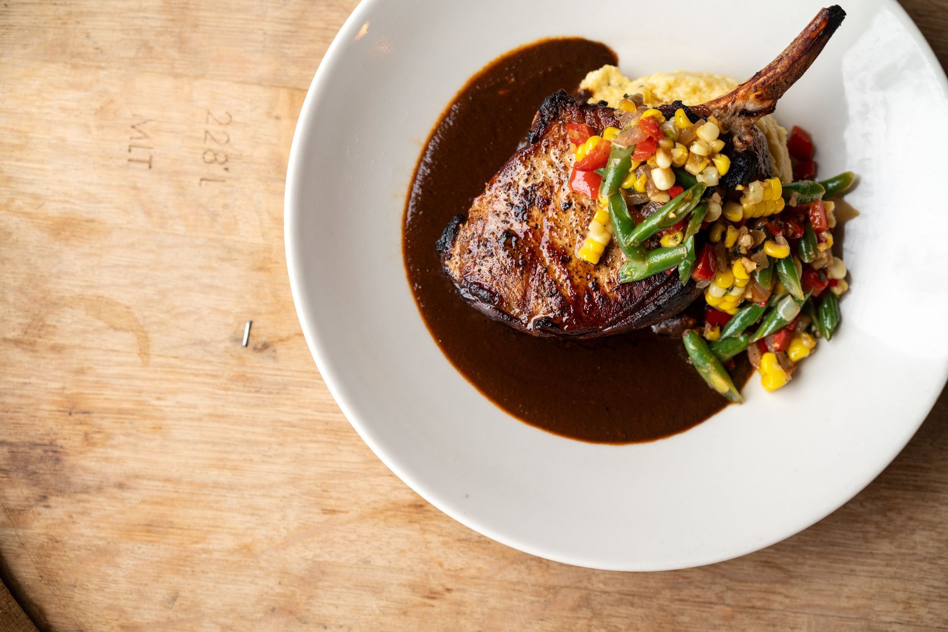 Grilled pork chop with sauce, corn, and green beans on a white plate, placed on a wooden table.