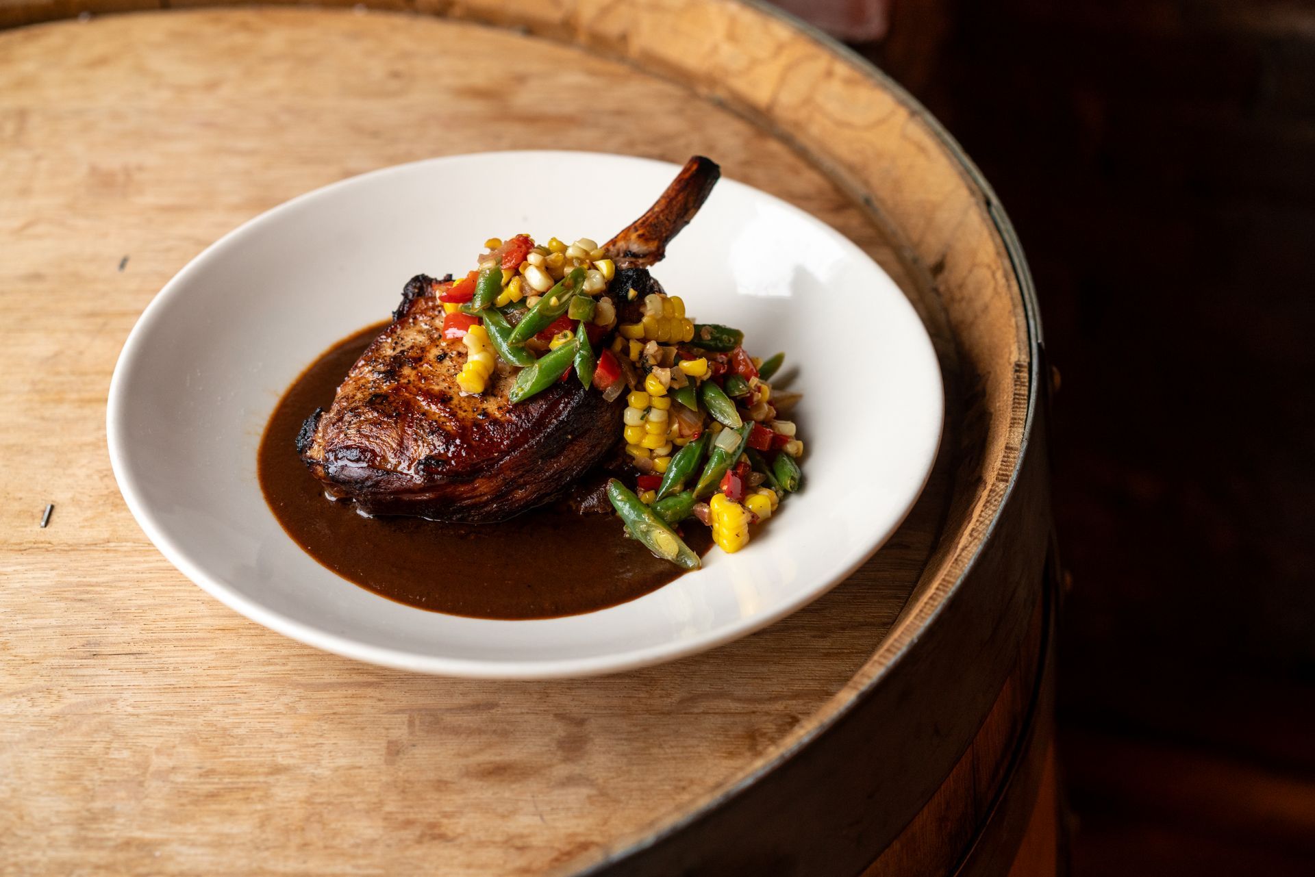 Pork chop with sauce and colorful relish on a white plate atop a wooden barrel.
