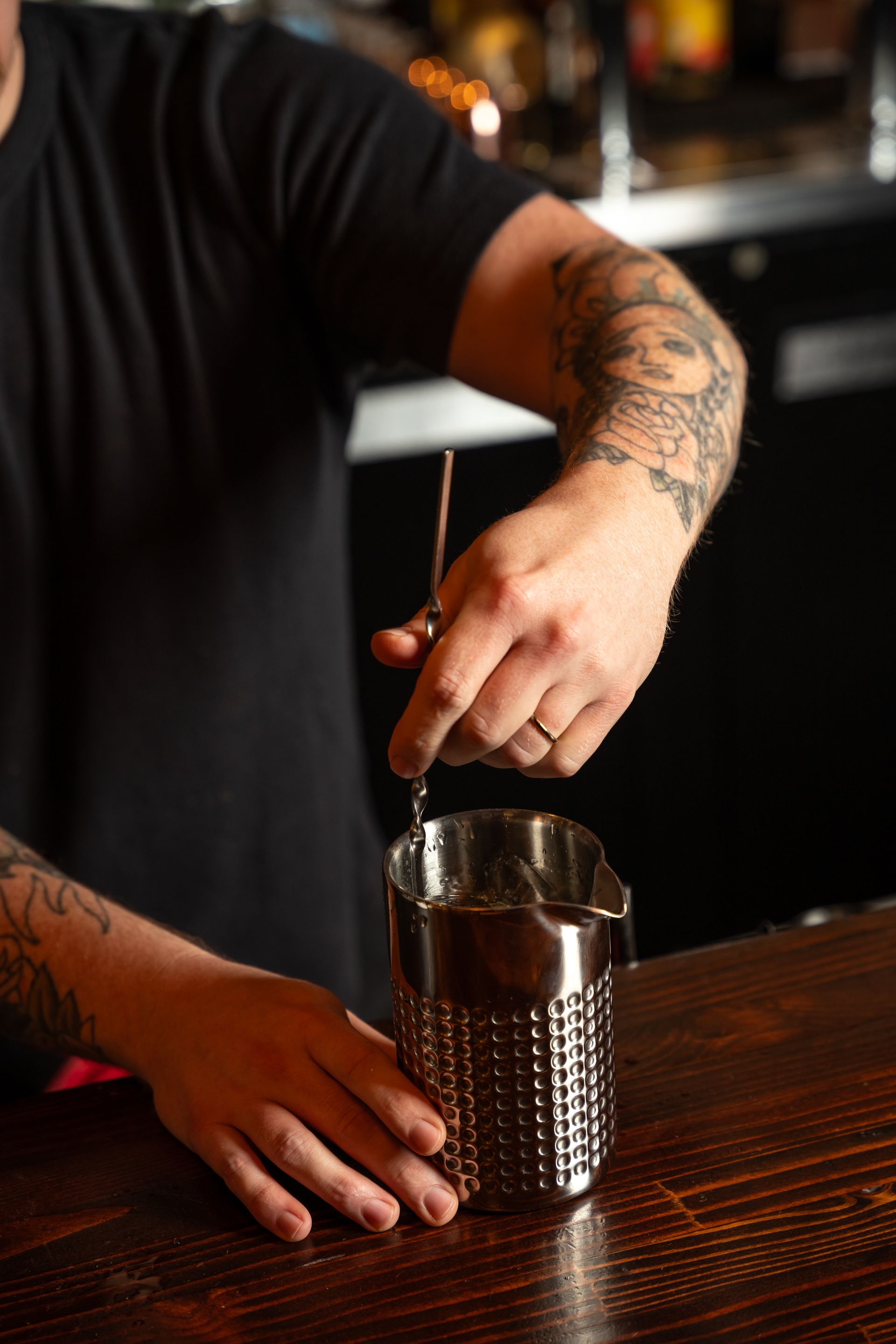 Bartender stirring a drink in a stainless steel shaker at a bar; tattoo visible on arm.