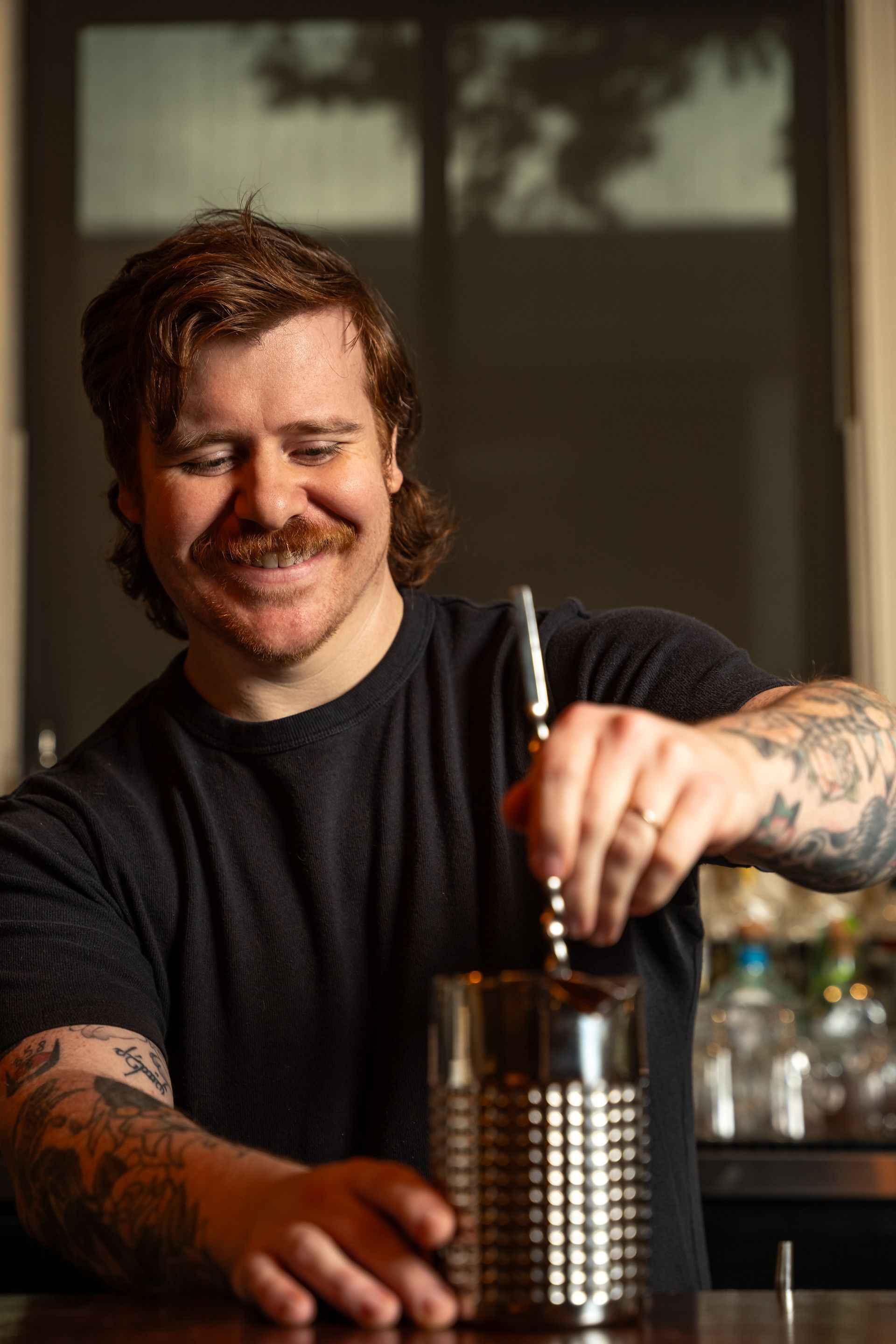 Bartender with red hair and mustache, stirring a drink, smiling. Dark shirt, tattoos on arms.