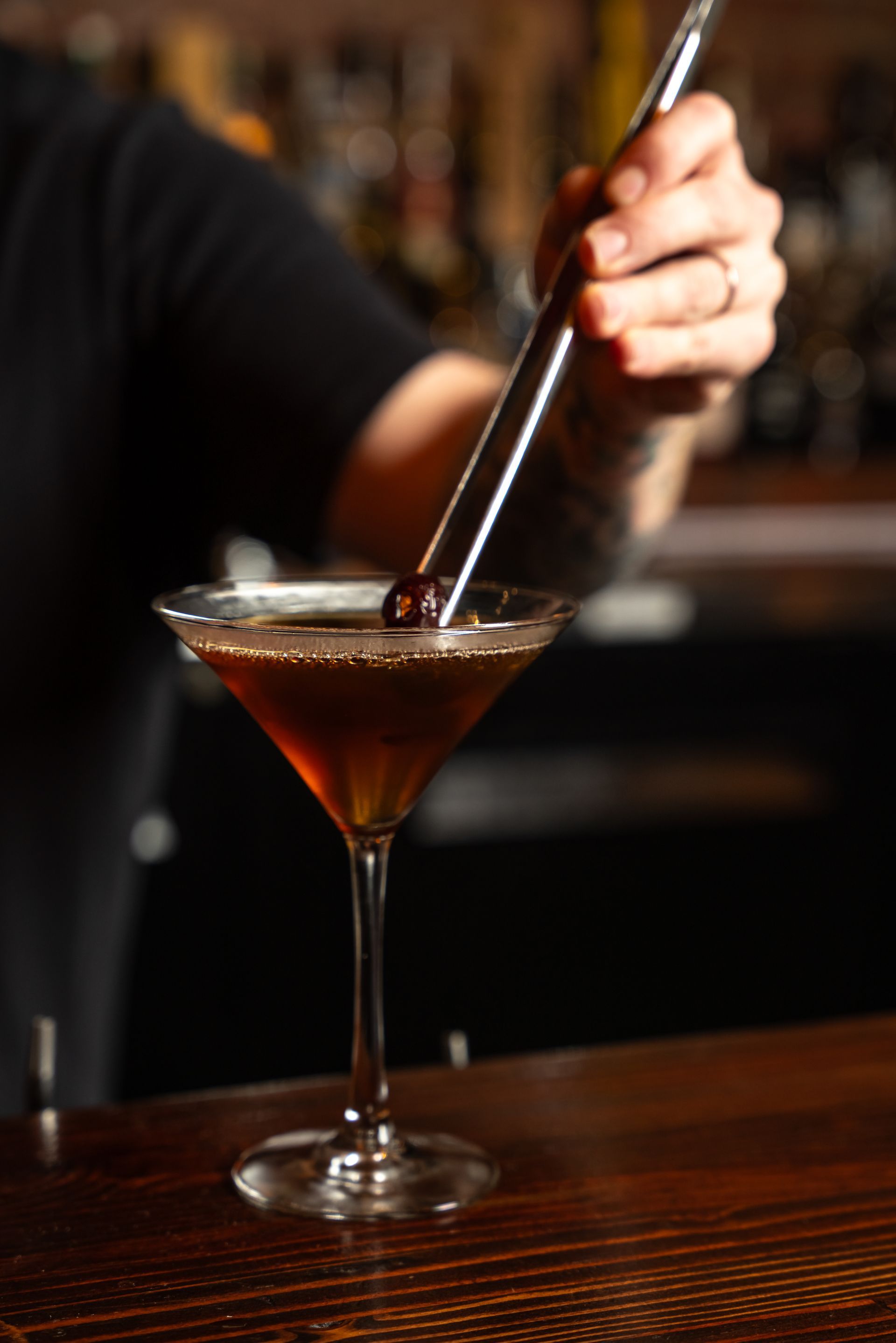 Bartender stirring a dark cocktail in a martini glass, garnished with a cherry.