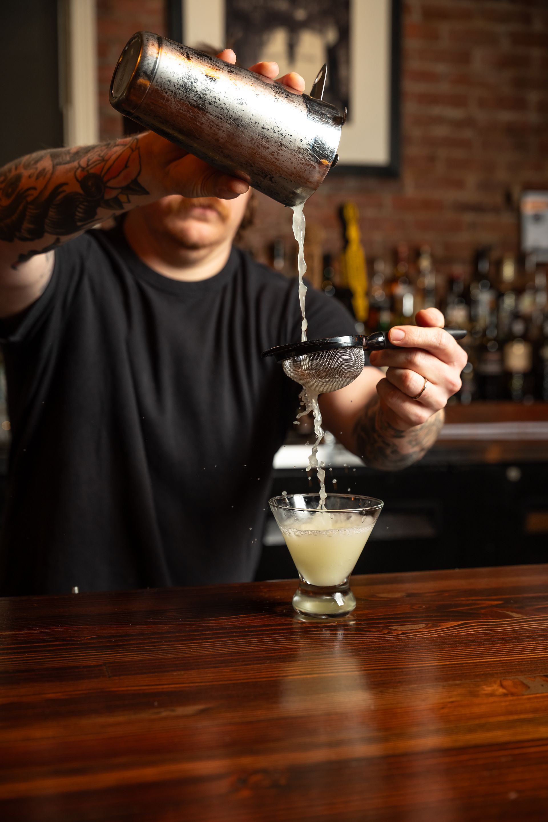 Bartender pouring a cocktail from a shaker into a glass, behind a wooden bar.