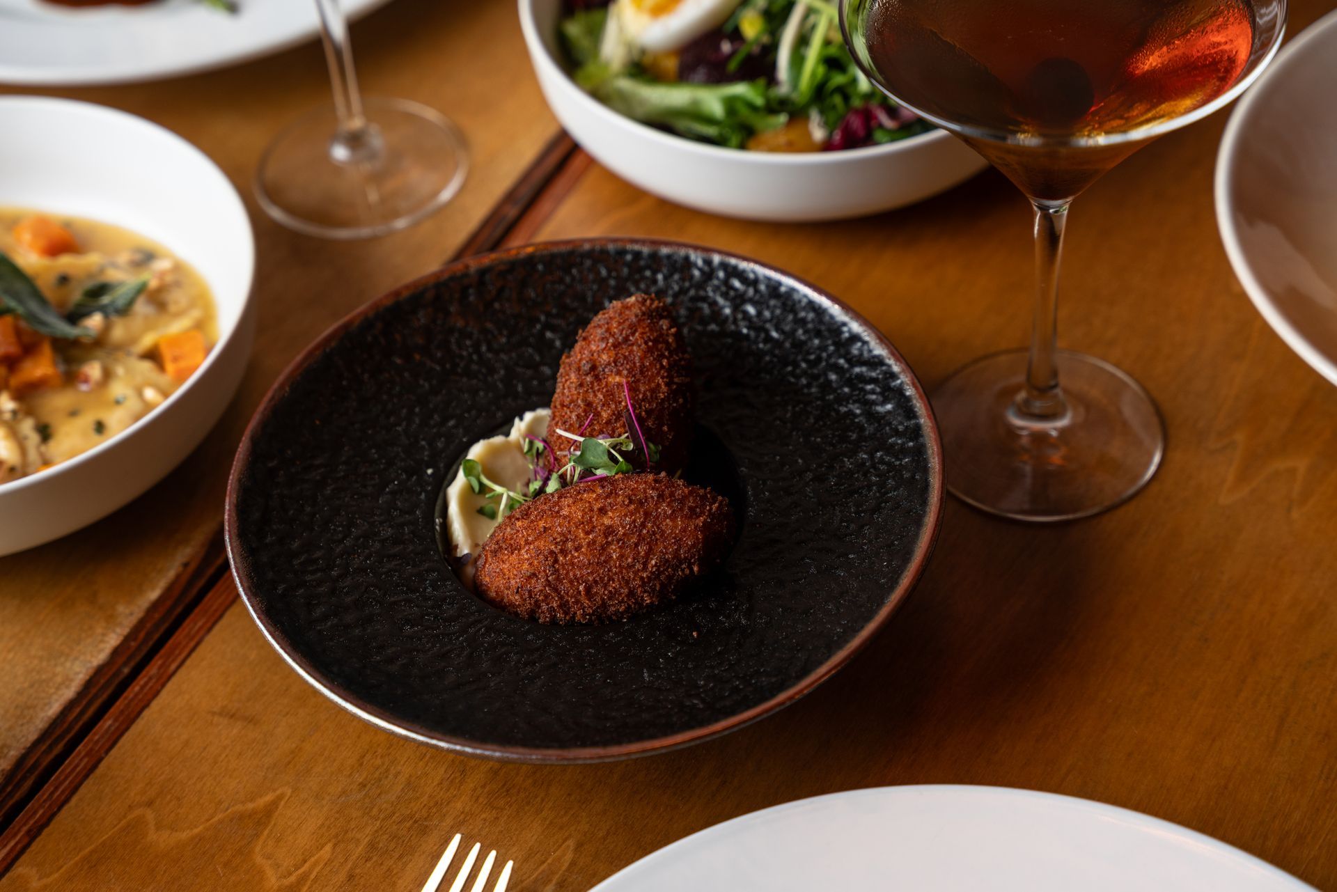 Two fried, oval food items on a black plate with white sauce and herbs, surrounded by other dishes and wine glasses on a wooden table.