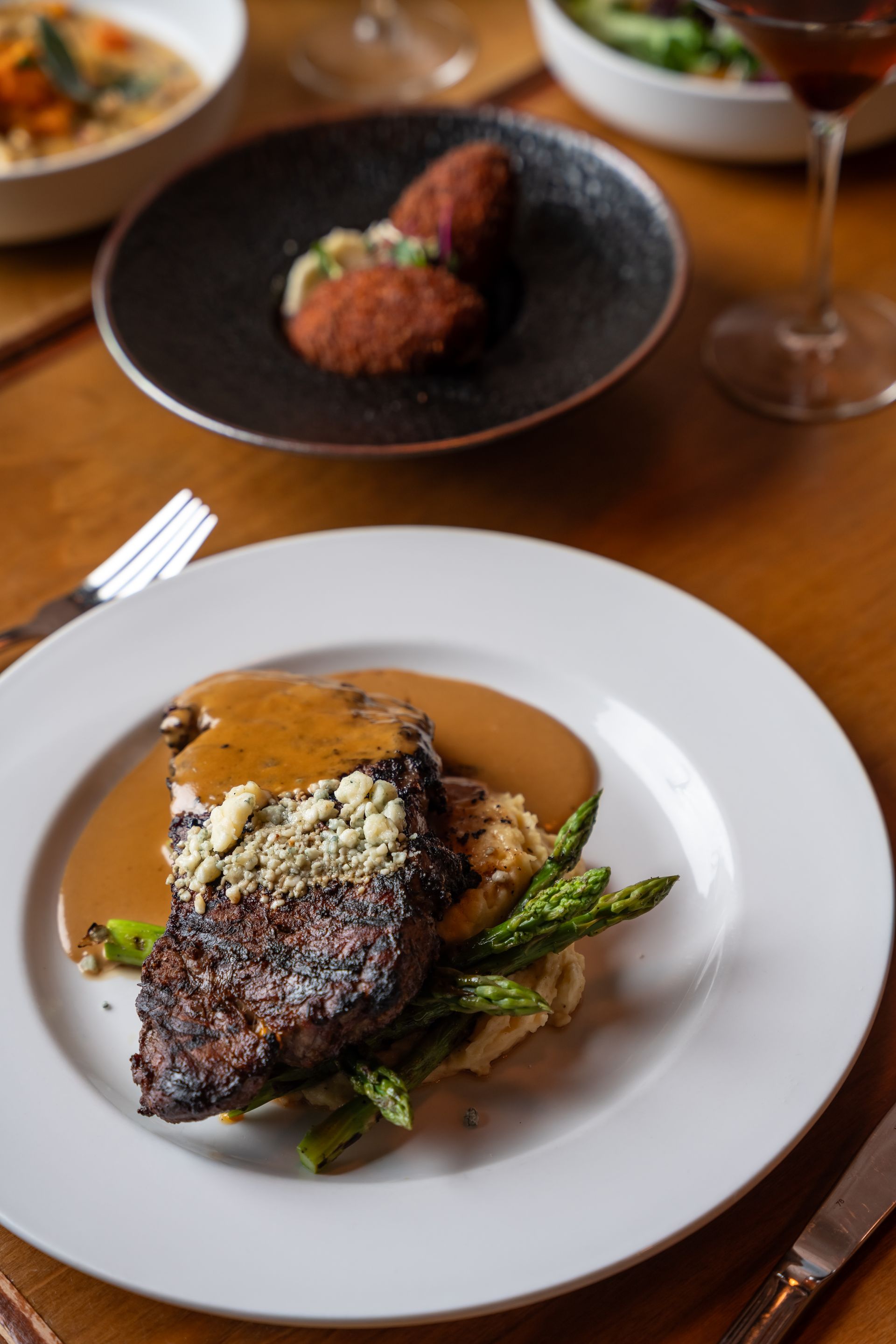 Steak with sauce, asparagus, and mashed potatoes on a white plate. Appetizers and a drink are in the background.