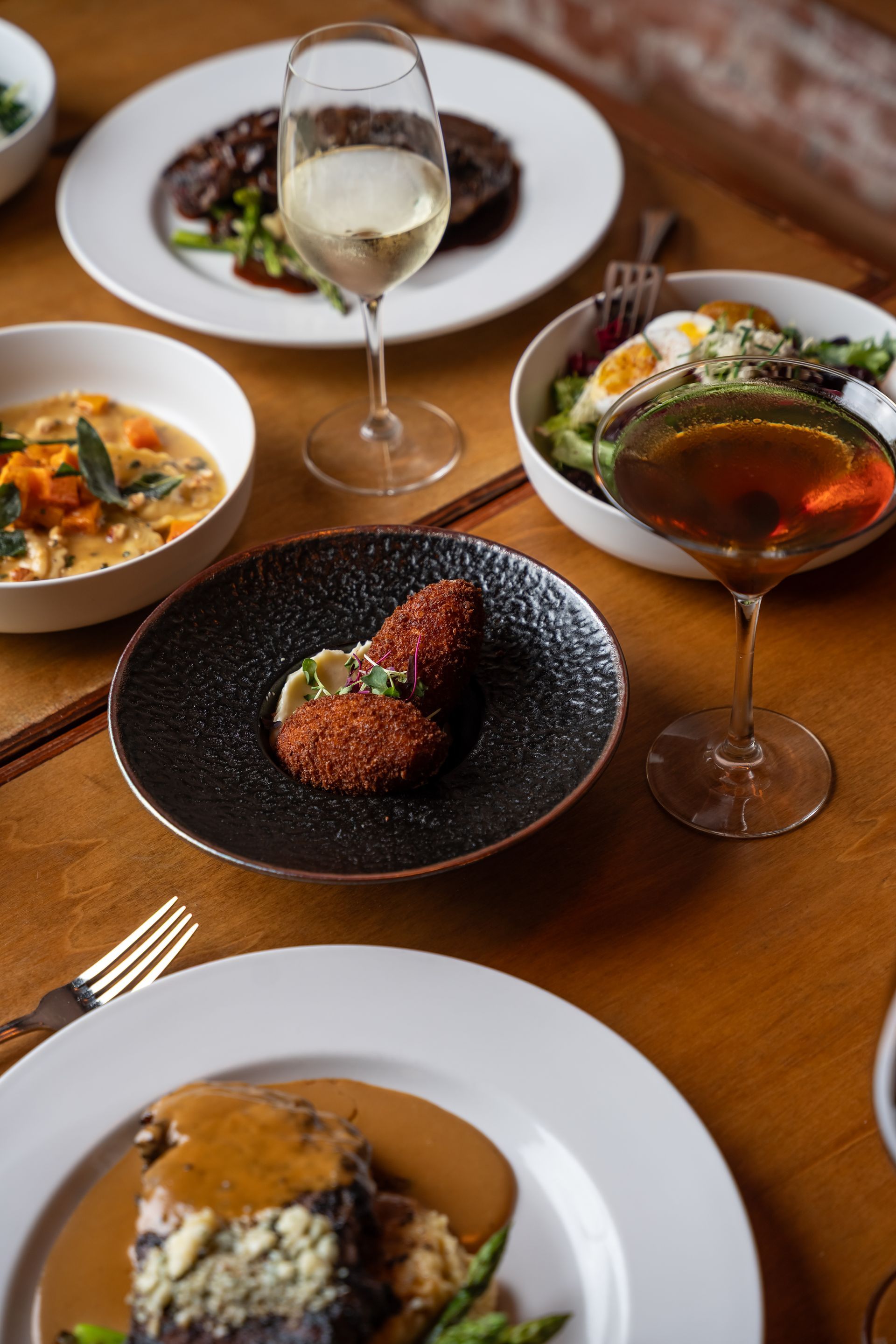 Various plates of food with drinks on a wooden table, likely a restaurant setting.