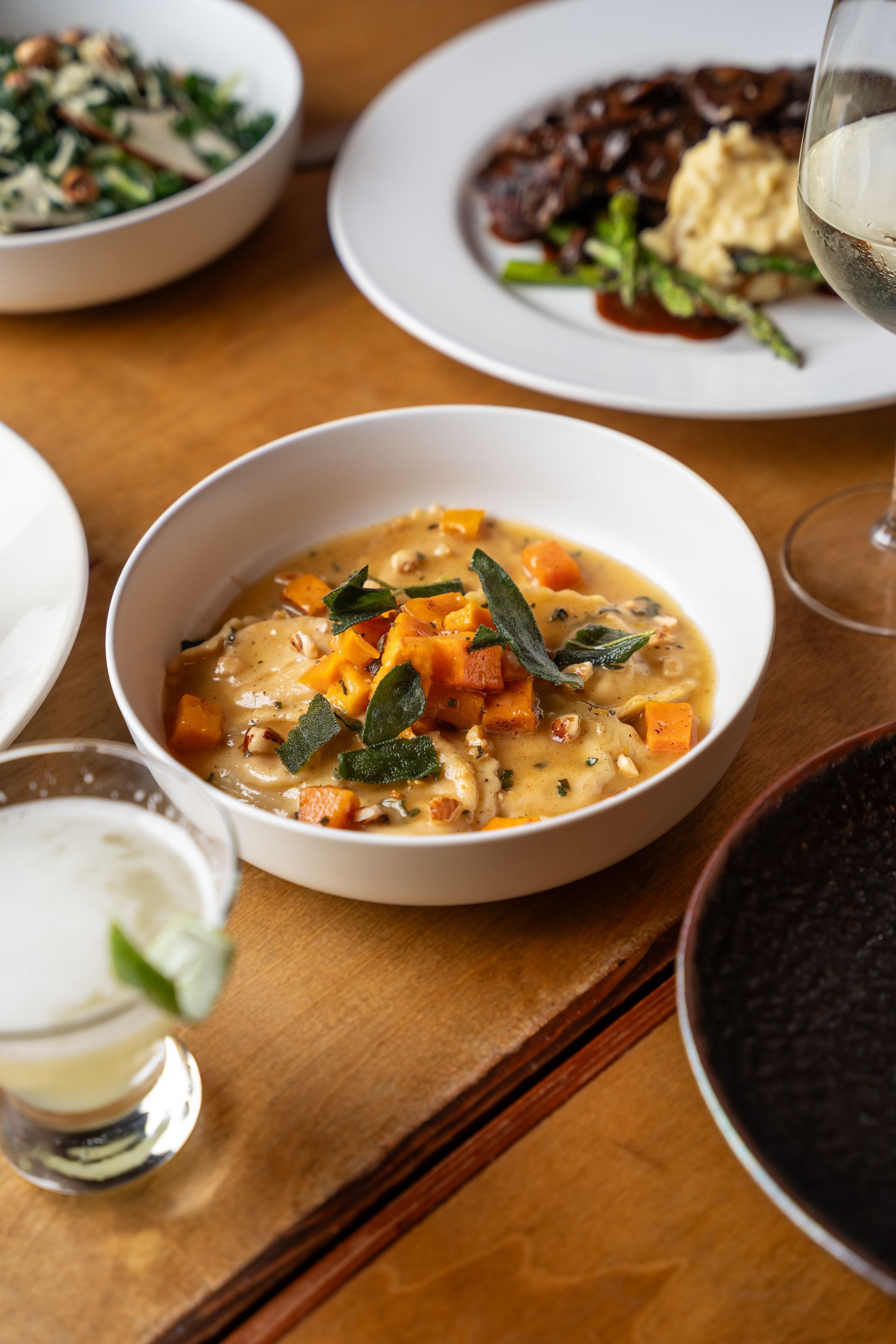 A bowl of ravioli with squash and sage, alongside other dishes and a cocktail on a wooden table.
