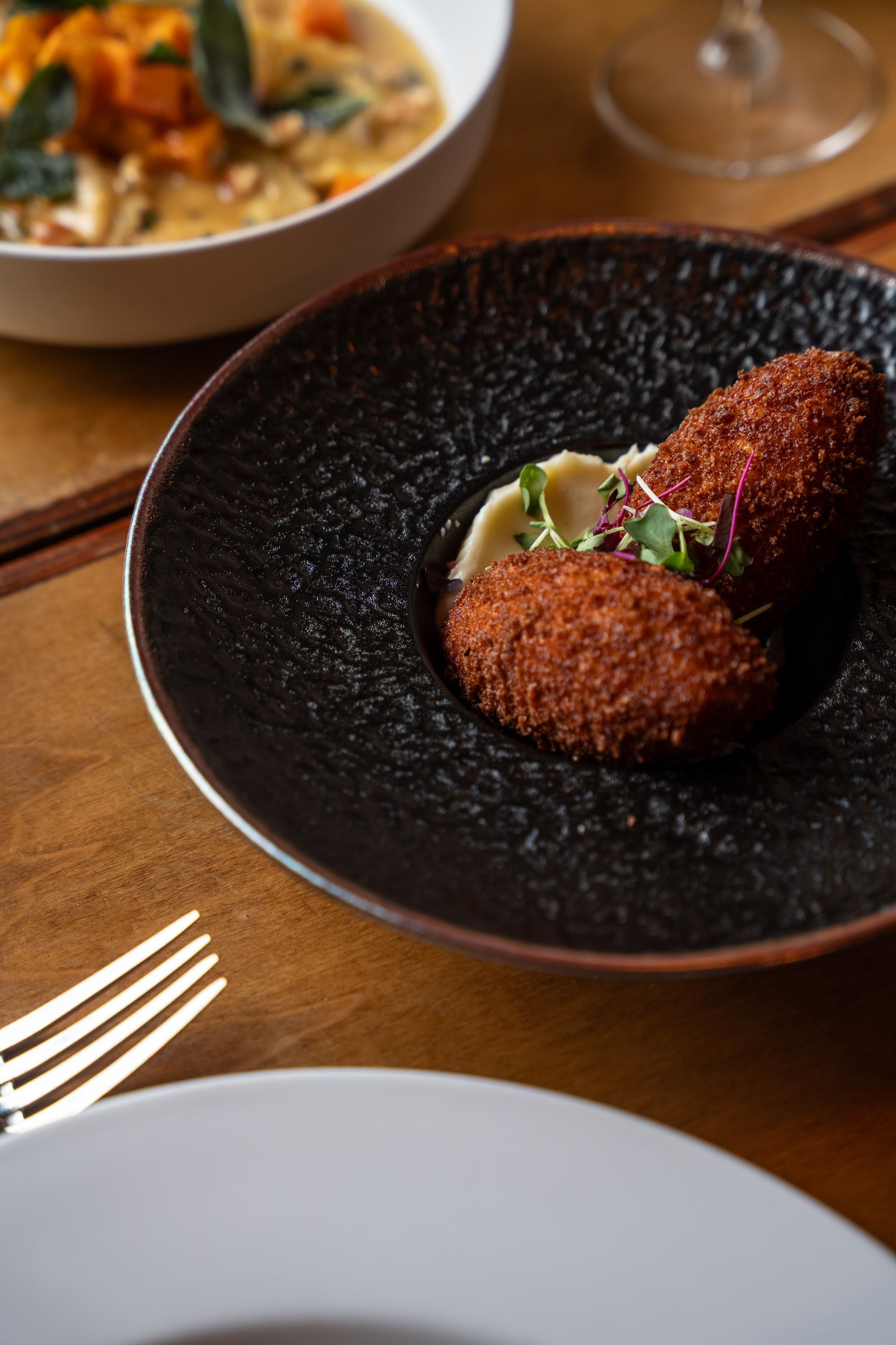 Two breaded, oblong croquettes on mashed potatoes, served on a black plate. A bowl of soup is partially visible in the background.