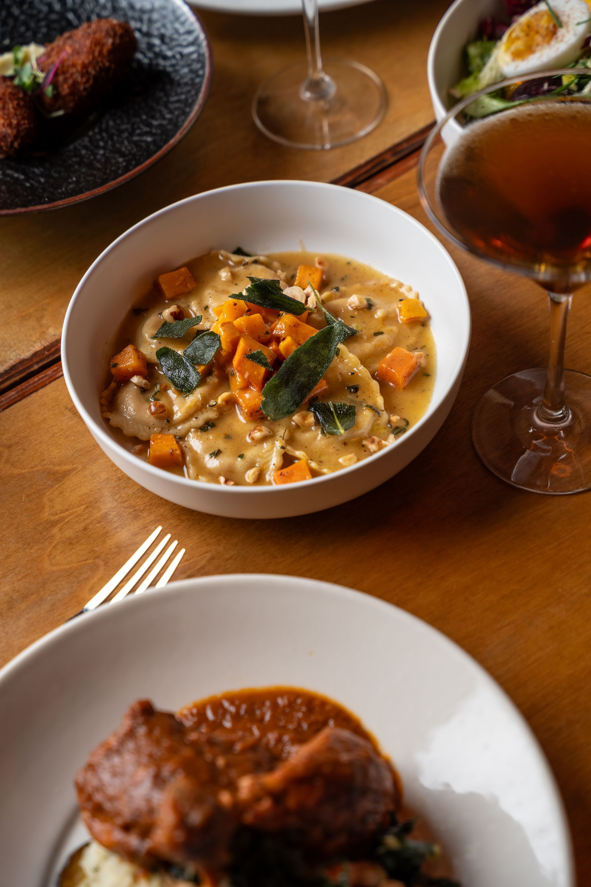 Various dishes on a brown table, ravioli, stew, salad, fried food, and wine.