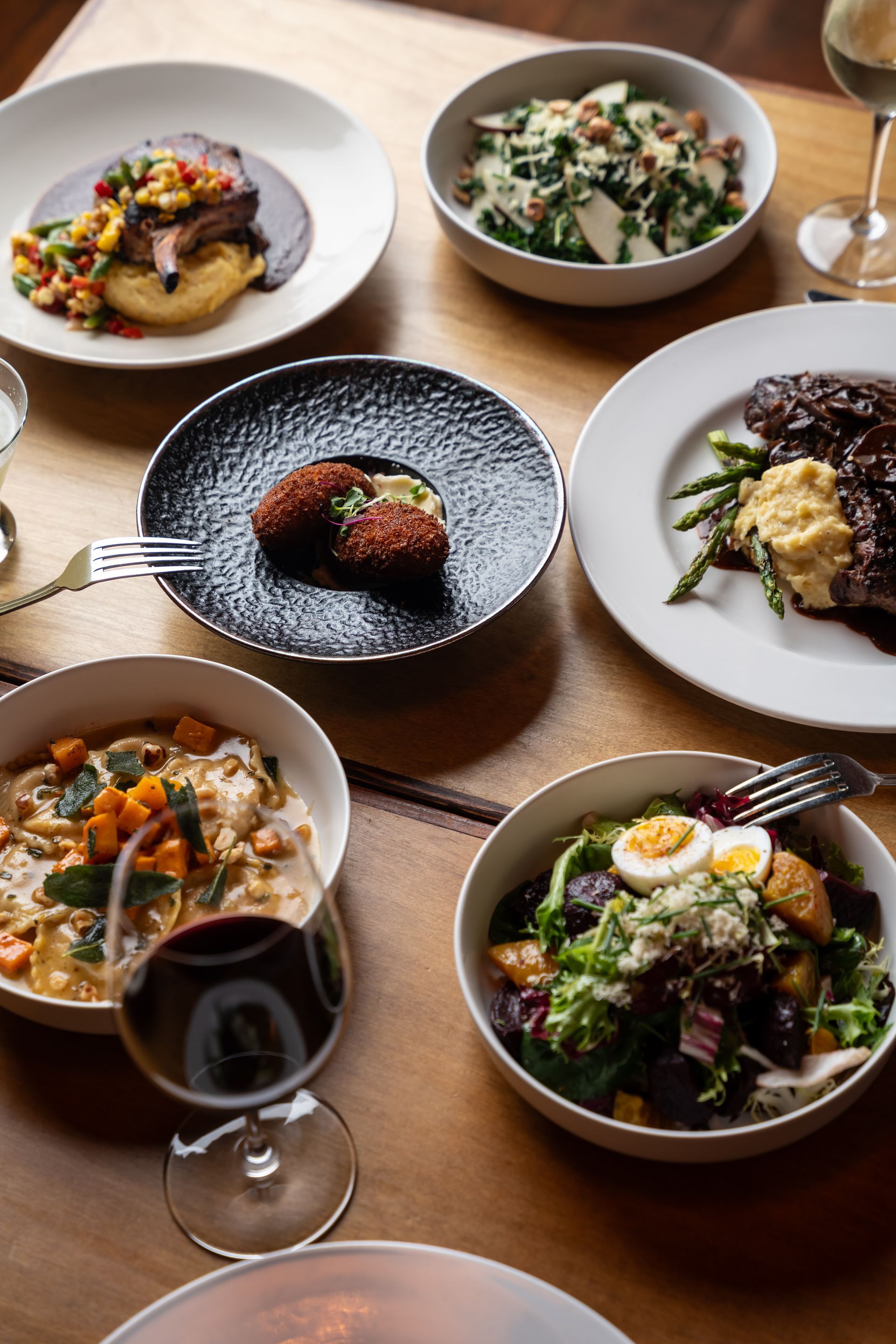 Assortment of plated dishes on a wooden table, including meat, salad, and pasta; wine glasses in view.