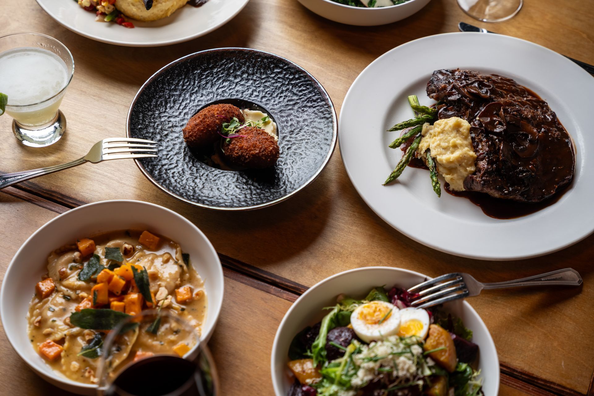 A table of various dishes, steak, ravioli, salad, and appetizers.