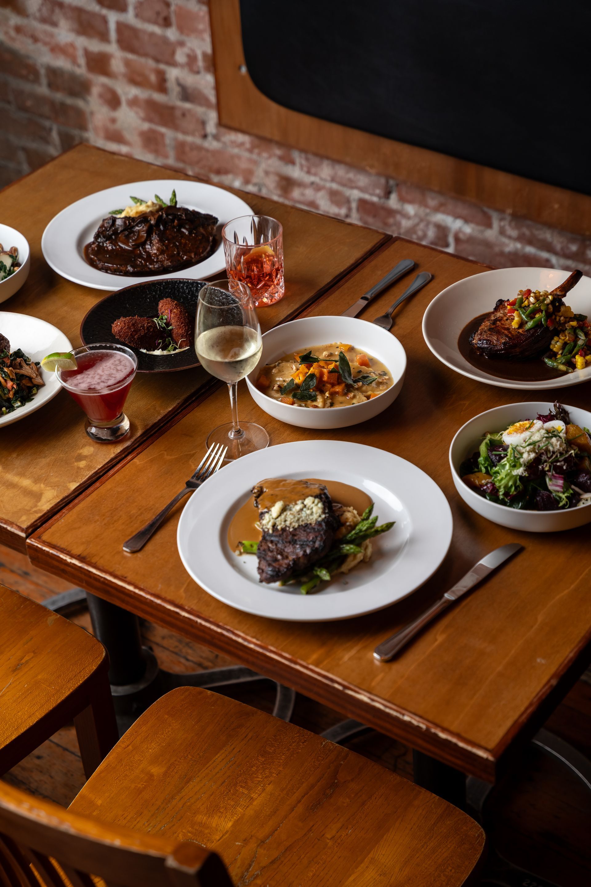 Wooden table set with various dishes, wine glasses, and cocktails, in a restaurant setting.