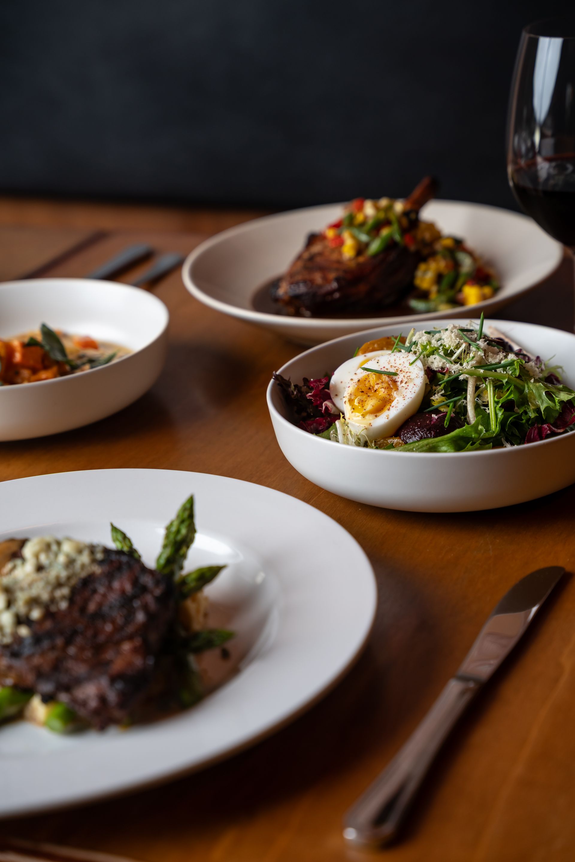 Several plates of gourmet food on a wooden table, including meat, salad, and a soup.