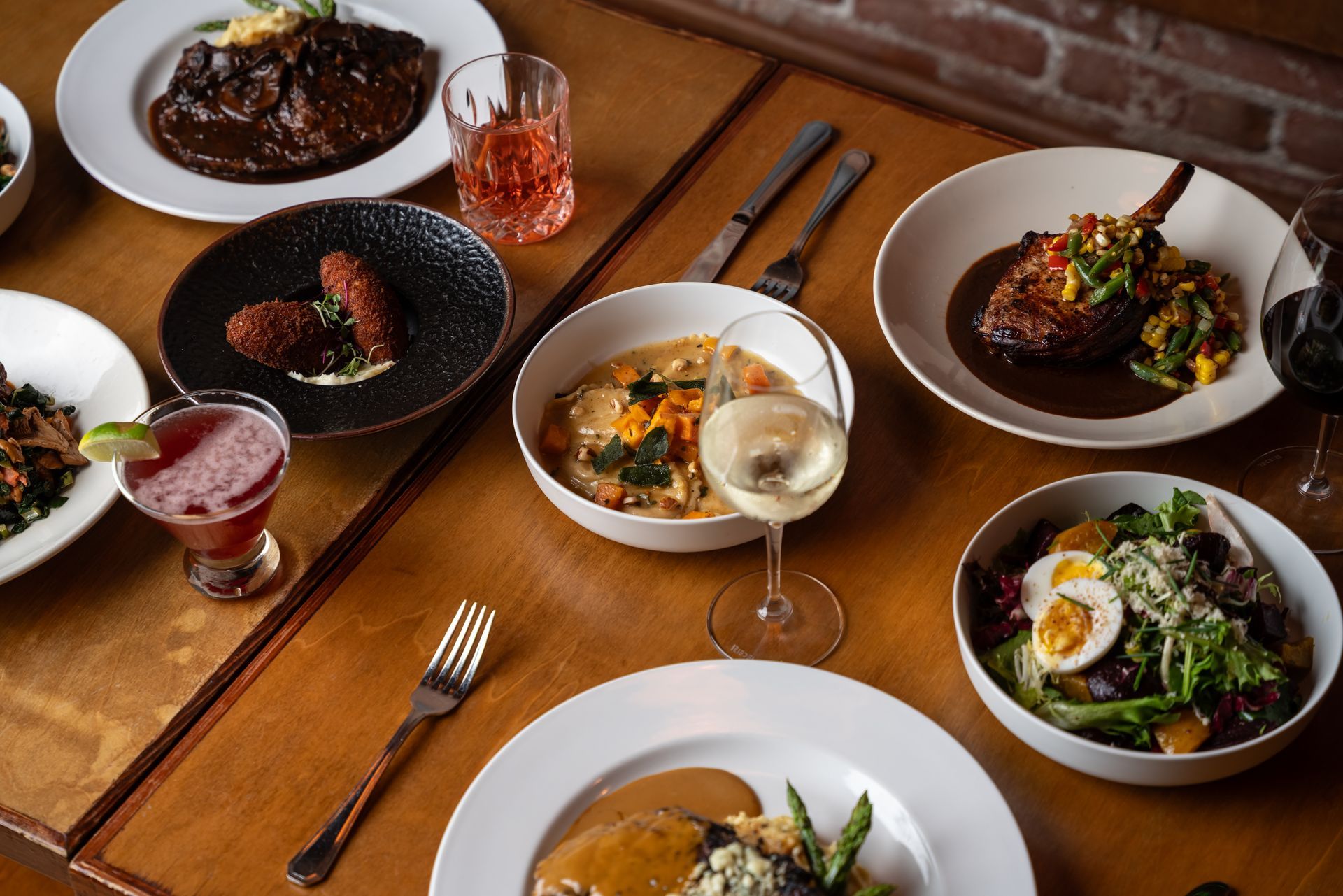 Table of various dishes, steak, salad, sides, cocktails, and wine.