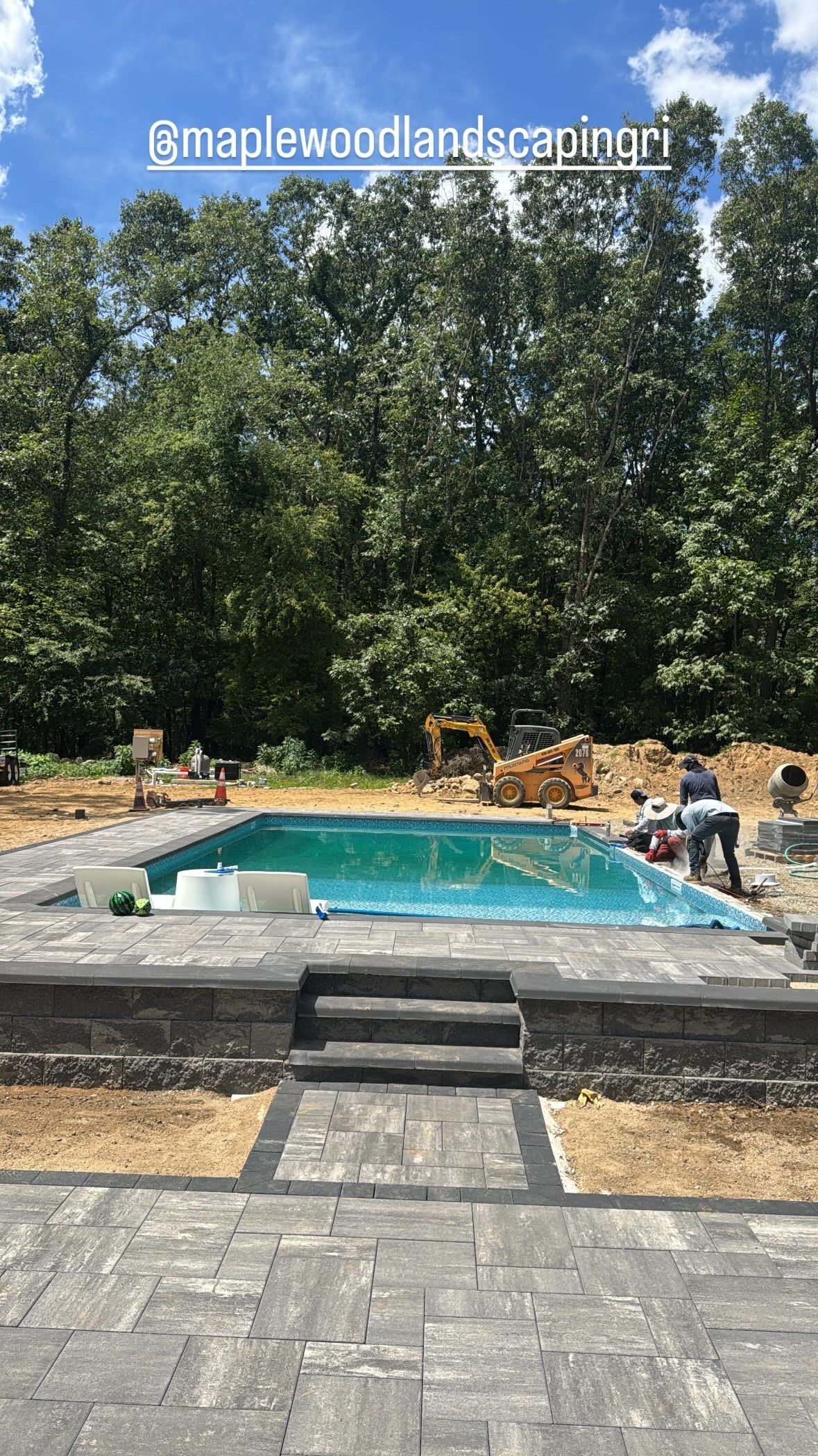 Rectangular pool with stone steps and surrounding lounge area, backed by tall green trees