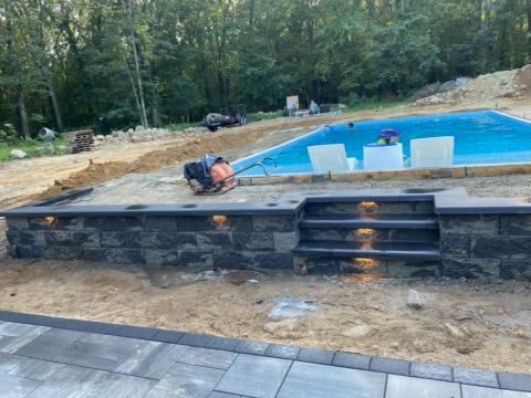Backyard pool under construction with stone retaining wall, steps, and stacked white lounge chairs