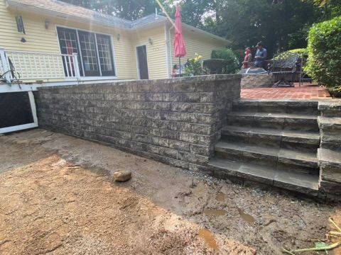 Stone retaining wall and steps leading to a patio beside a light-colored house and sandy yard.