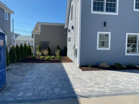 Paved patio and walkway between blue-gray houses with landscaped shrubs and a fenced yard.