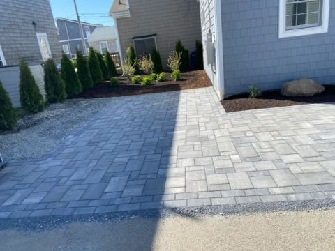 Paved backyard patio and walkway with gray pavers beside a house and landscaped shrubs