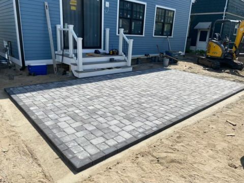 Stone paver patio under construction beside a blue house, with steps and a small excavator nearby