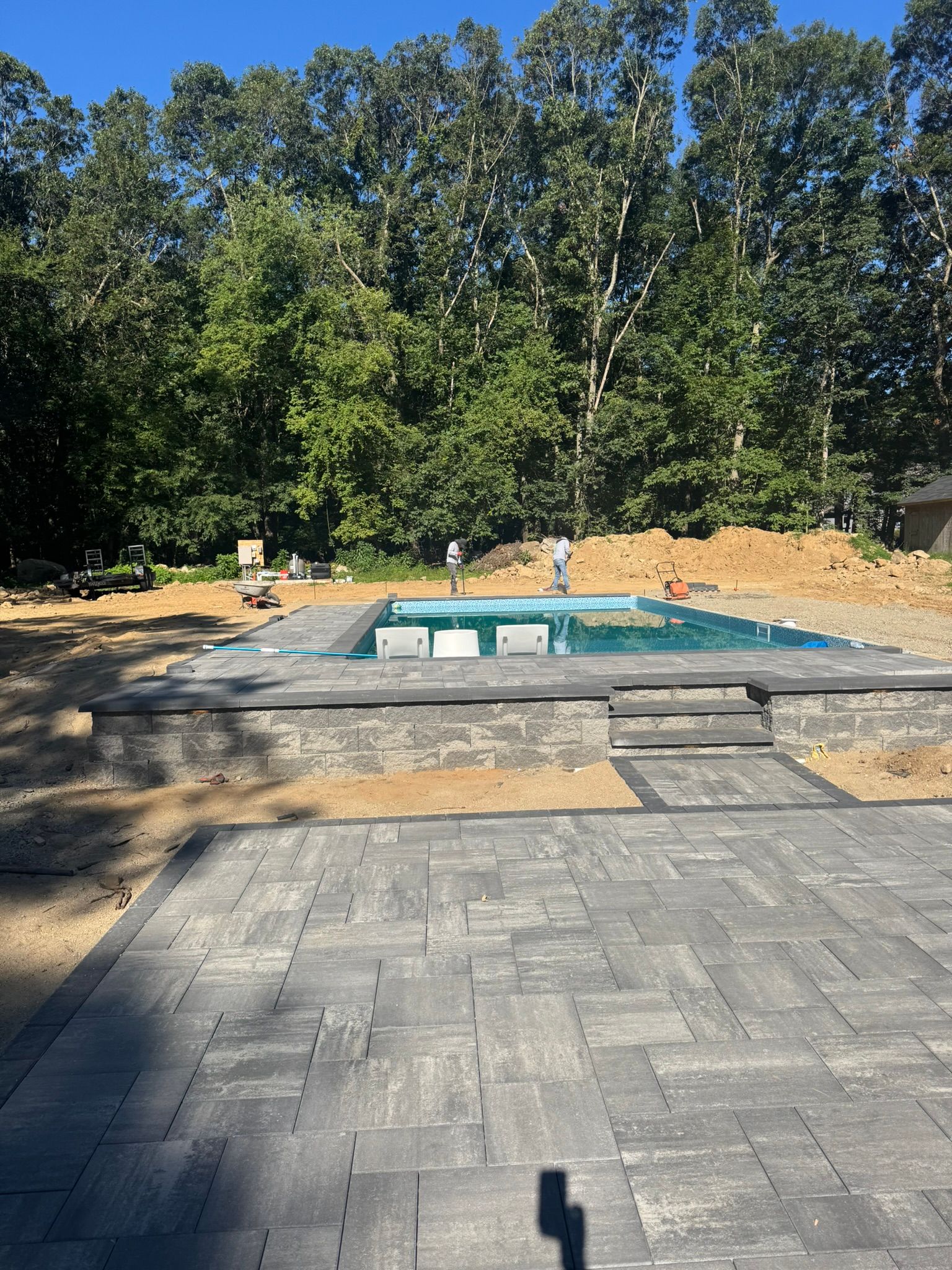 Backyard patio facing a rectangular pool under construction, with pavers and trees in the background