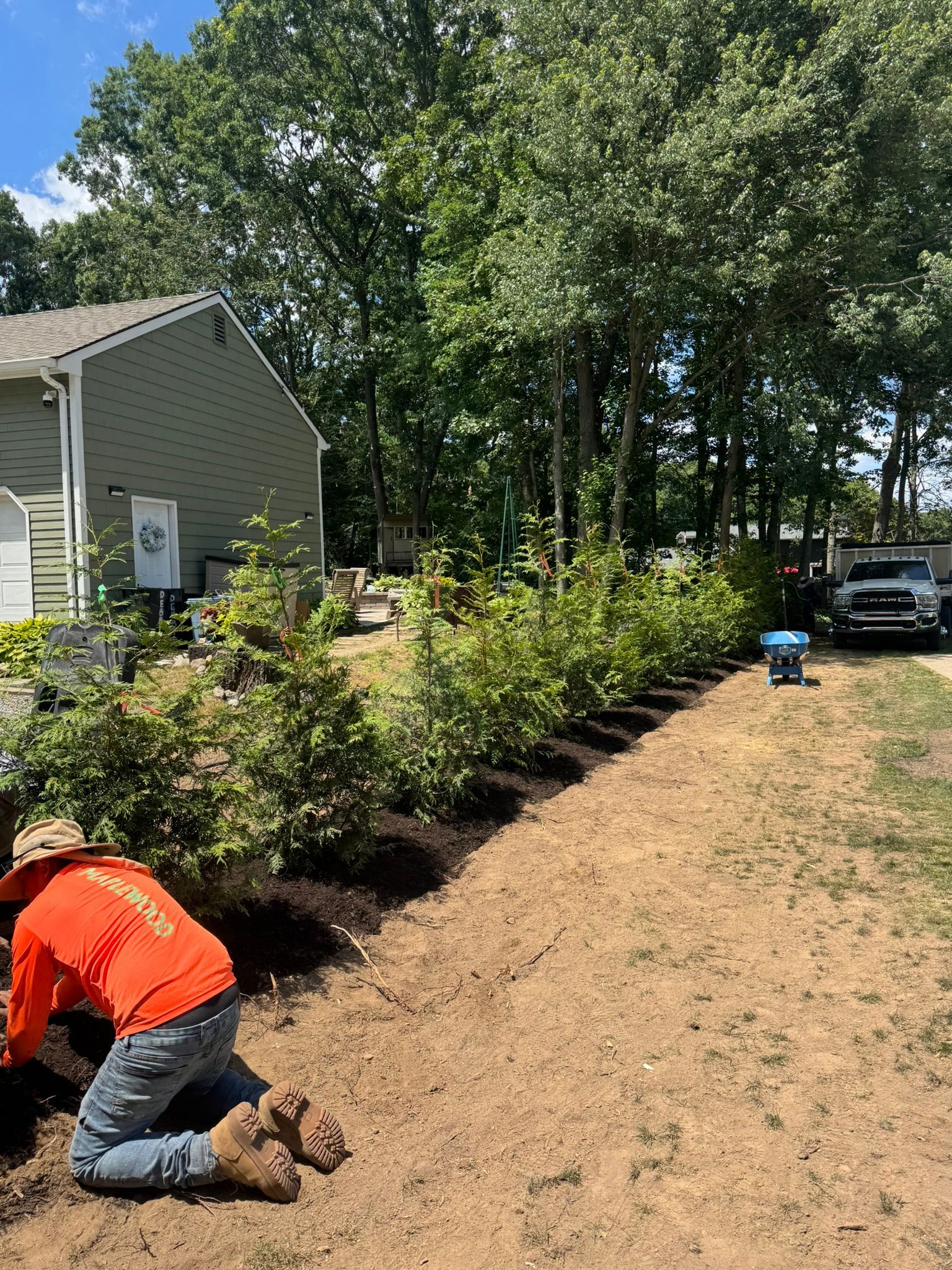 Worker in orange jacket kneels in mulch beside a house, landscaping a garden bed near a driveway.