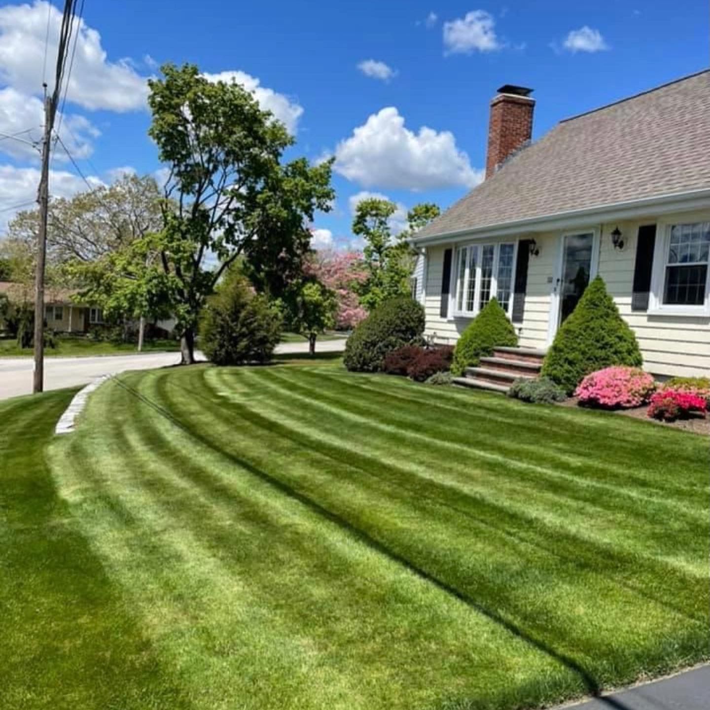 A bright blue sky over a house with light yellow siding and a manicured lawn with prominent, curved mowing stripes.
