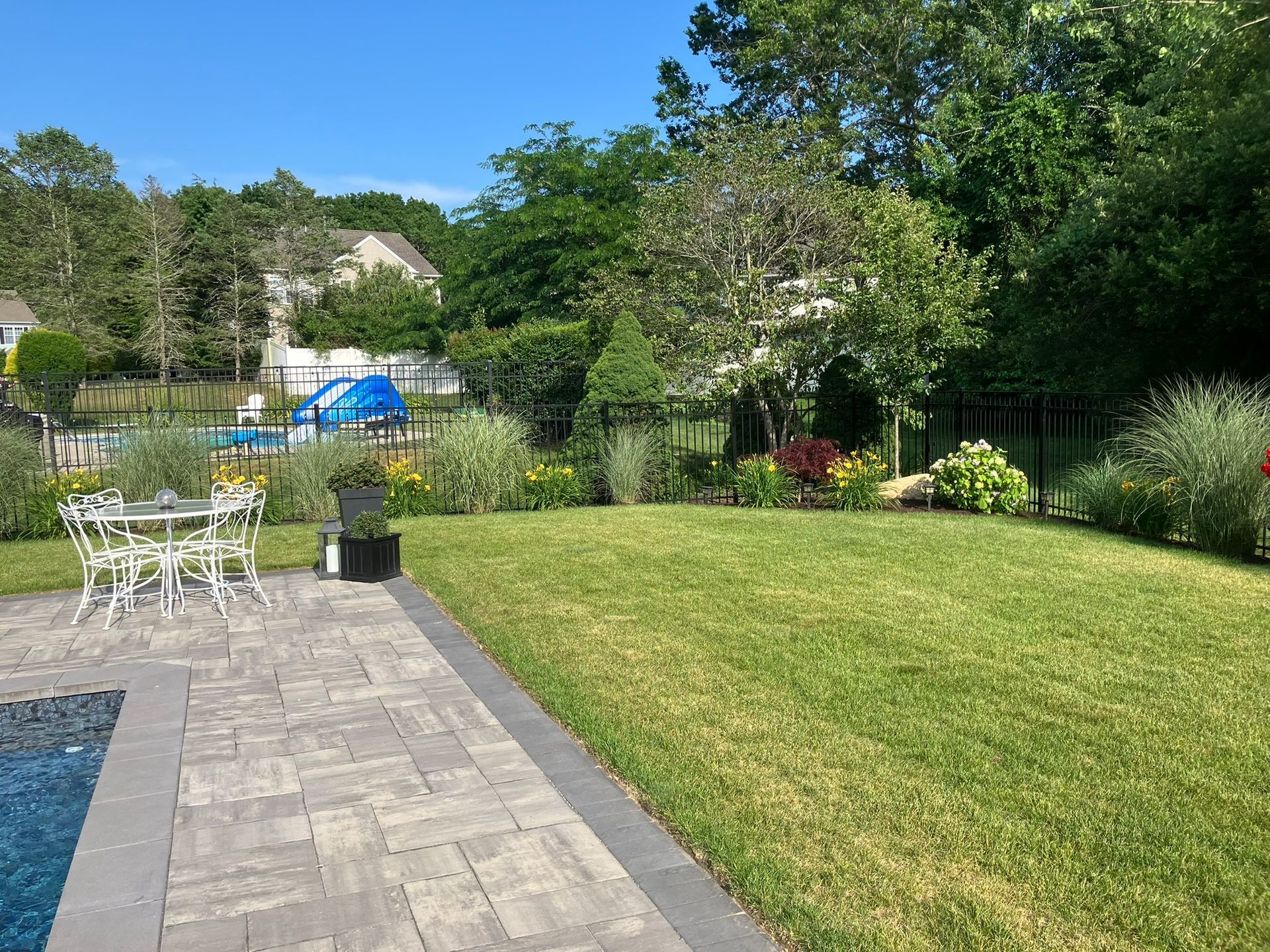 A sunny backyard with a pool, stone patio, white metal dining set, and lush green lawn bordered by garden beds and trees.