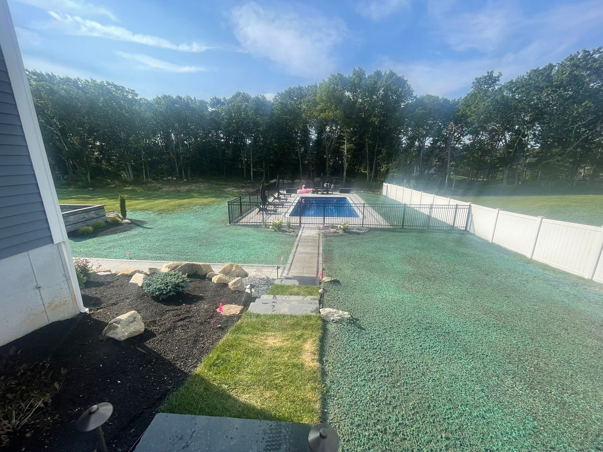 A backyard with a small rectangular pool, a white fence, and a green-tinted lawn area, viewed from a patio.