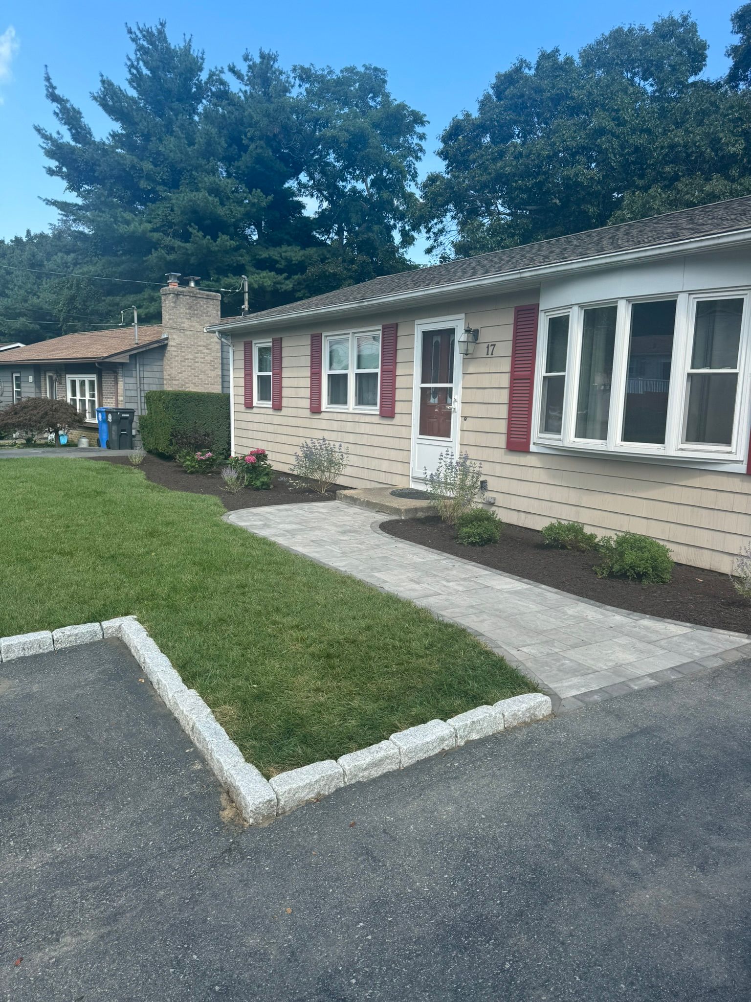 A tan, single-story house with a brick facade, red shutters, and a newly paved light-gray stone walkway and lawn border.