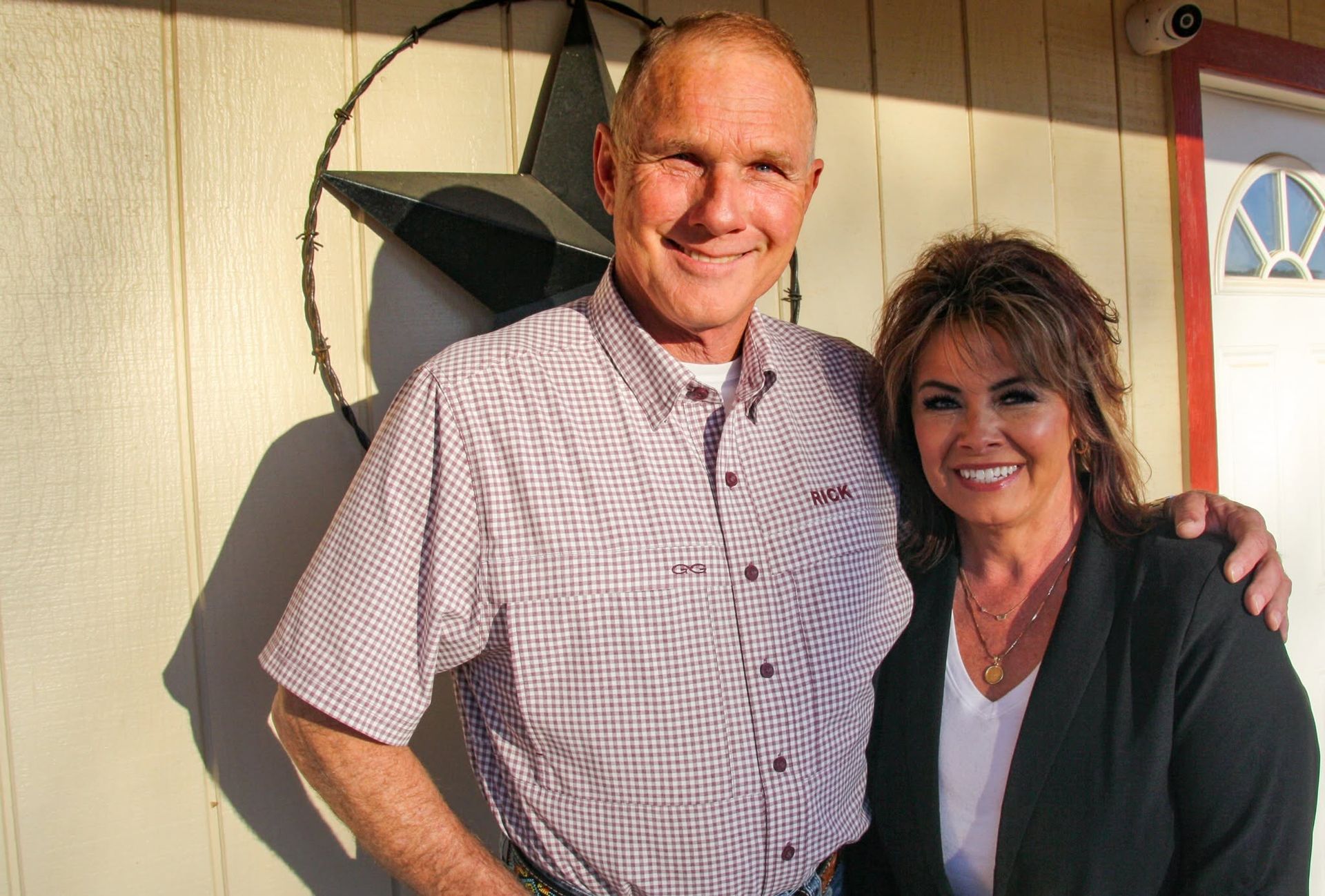 Two people smiling and posing outdoors in front of a house door, one with an arm around the other.