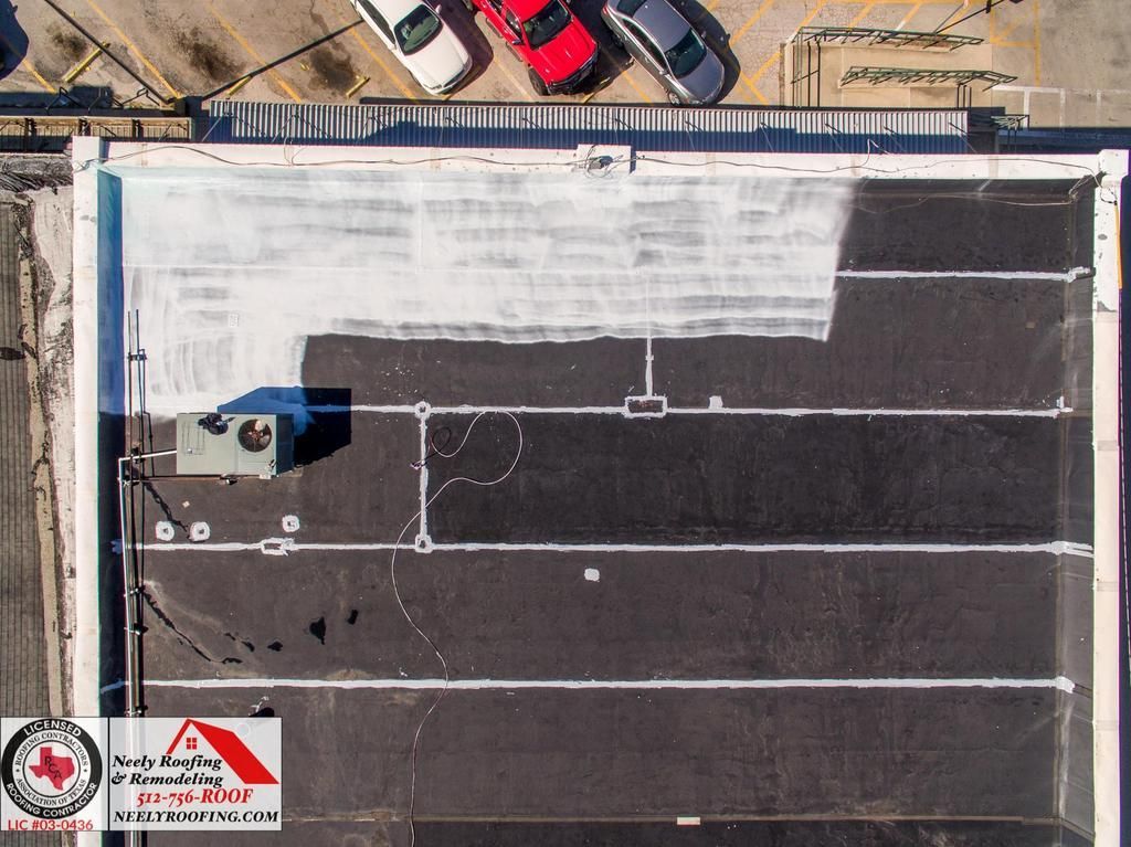 Aerial view of a flat rooftop with dark sections, white coating, and a small HVAC unit near the edge.