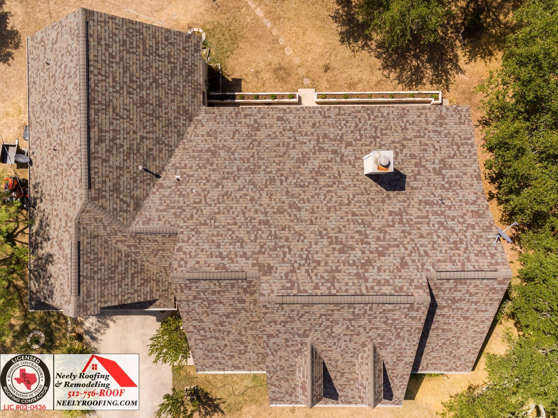 Aerial view of a house with a gray shingle roof and small chimney, surrounded by trees and dry grass.