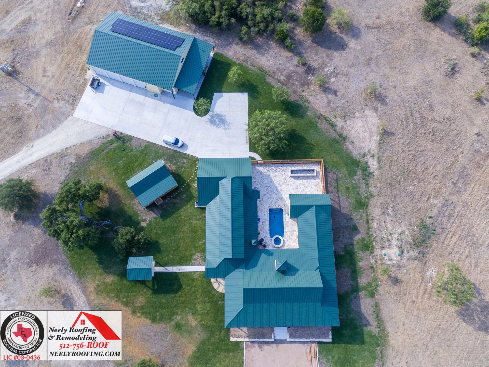 Aerial view of a house with teal roofs, a driveway, and small outbuildings on a dry lot.