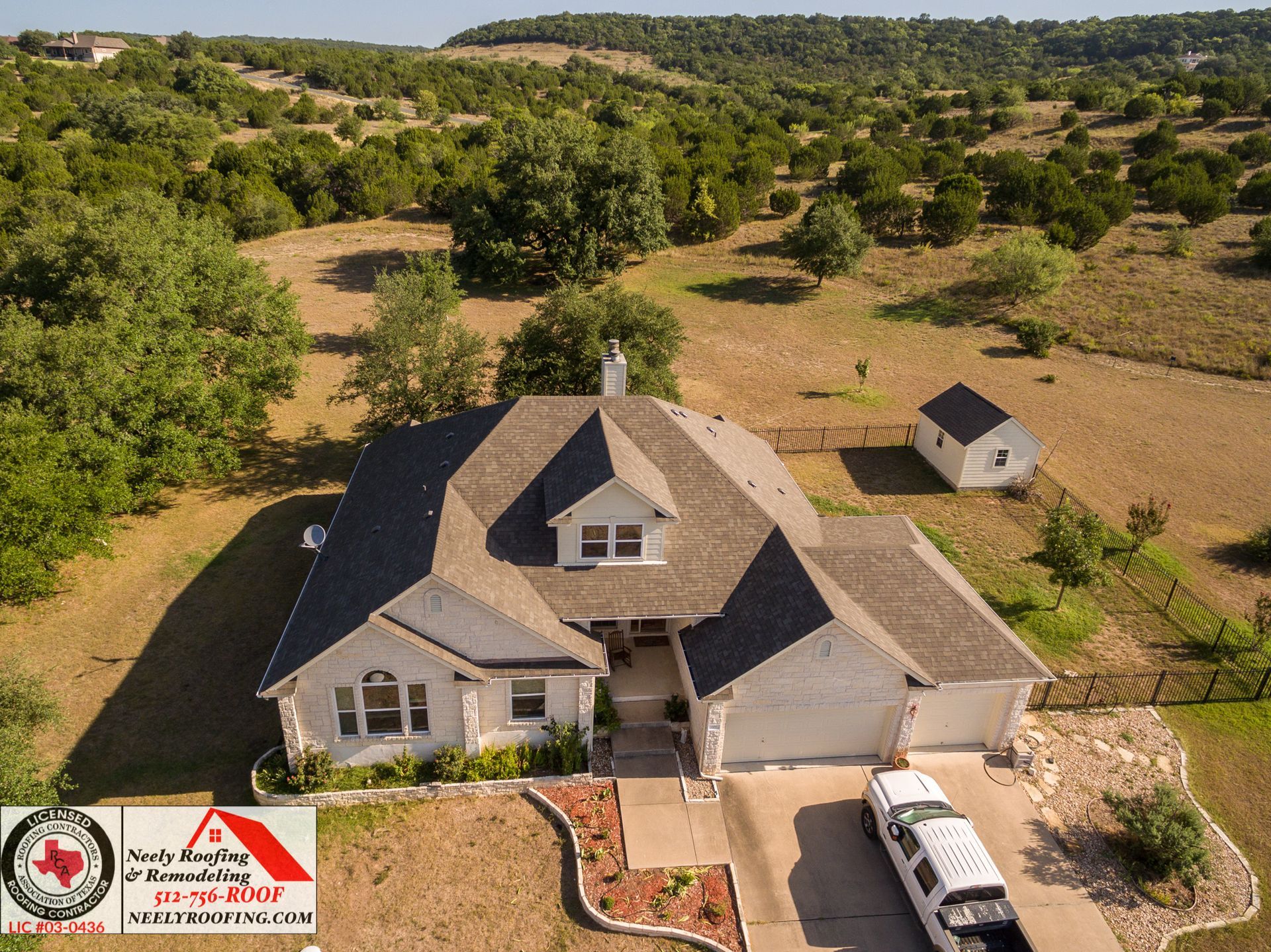 Aerial view of a gray house with a driveway and surrounded by trees and open land.