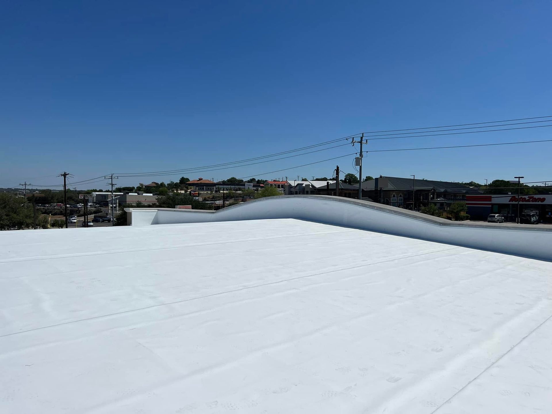 White rooftop with a low parapet under a clear blue sky, overlooking distant buildings and power lines