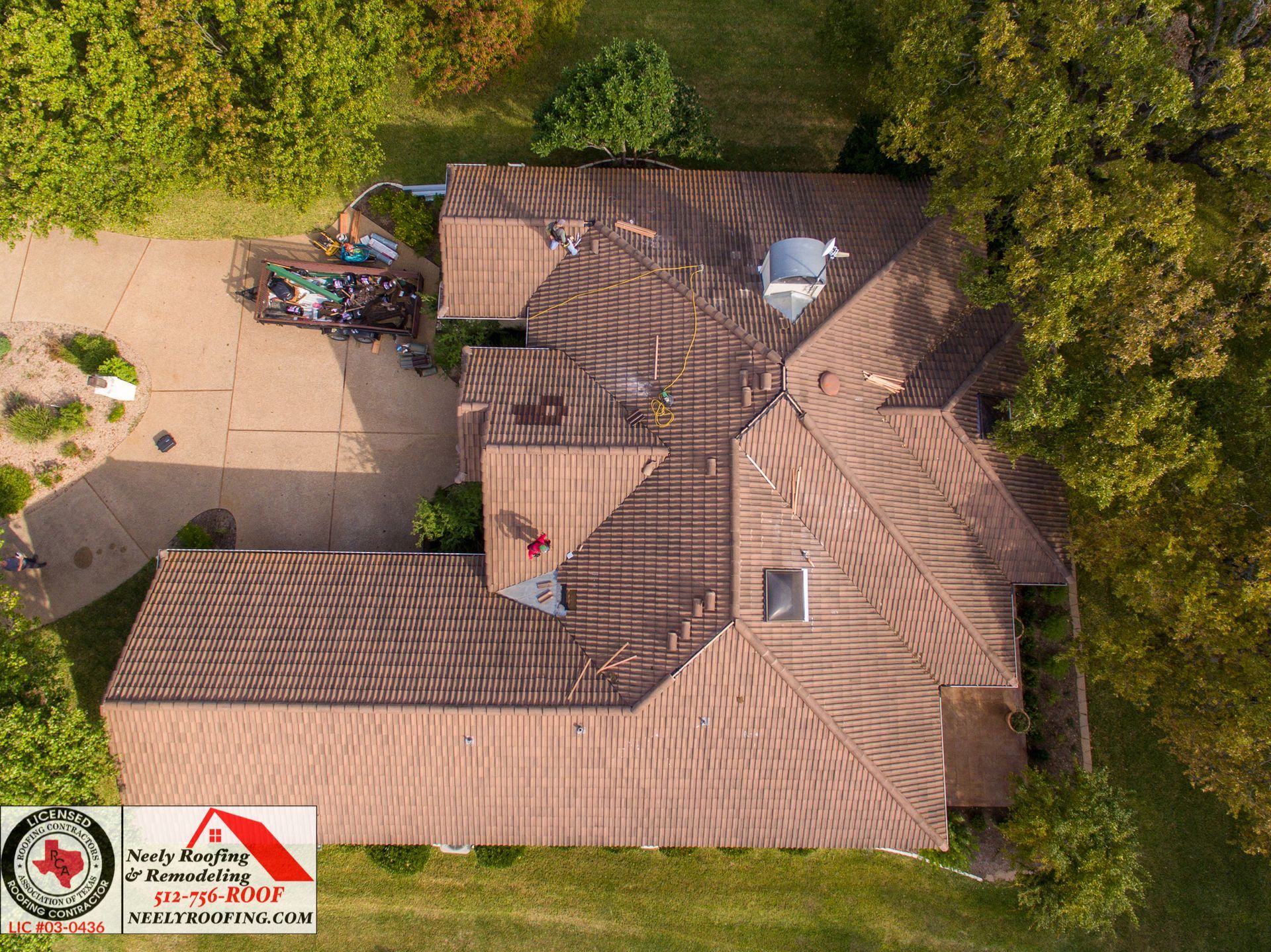 Aerial view of a brown-roofed house beside a driveway and trees, with a small image watermark in the corner
