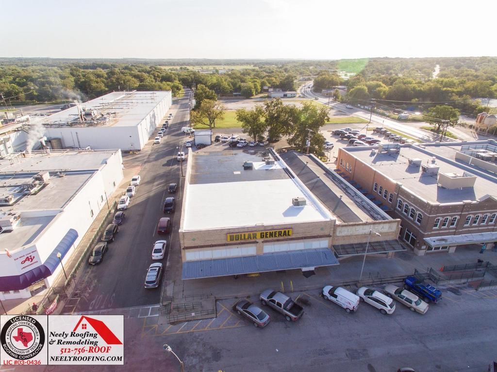Aerial view of a small commercial strip with parked cars, low-rise buildings, and trees in the background