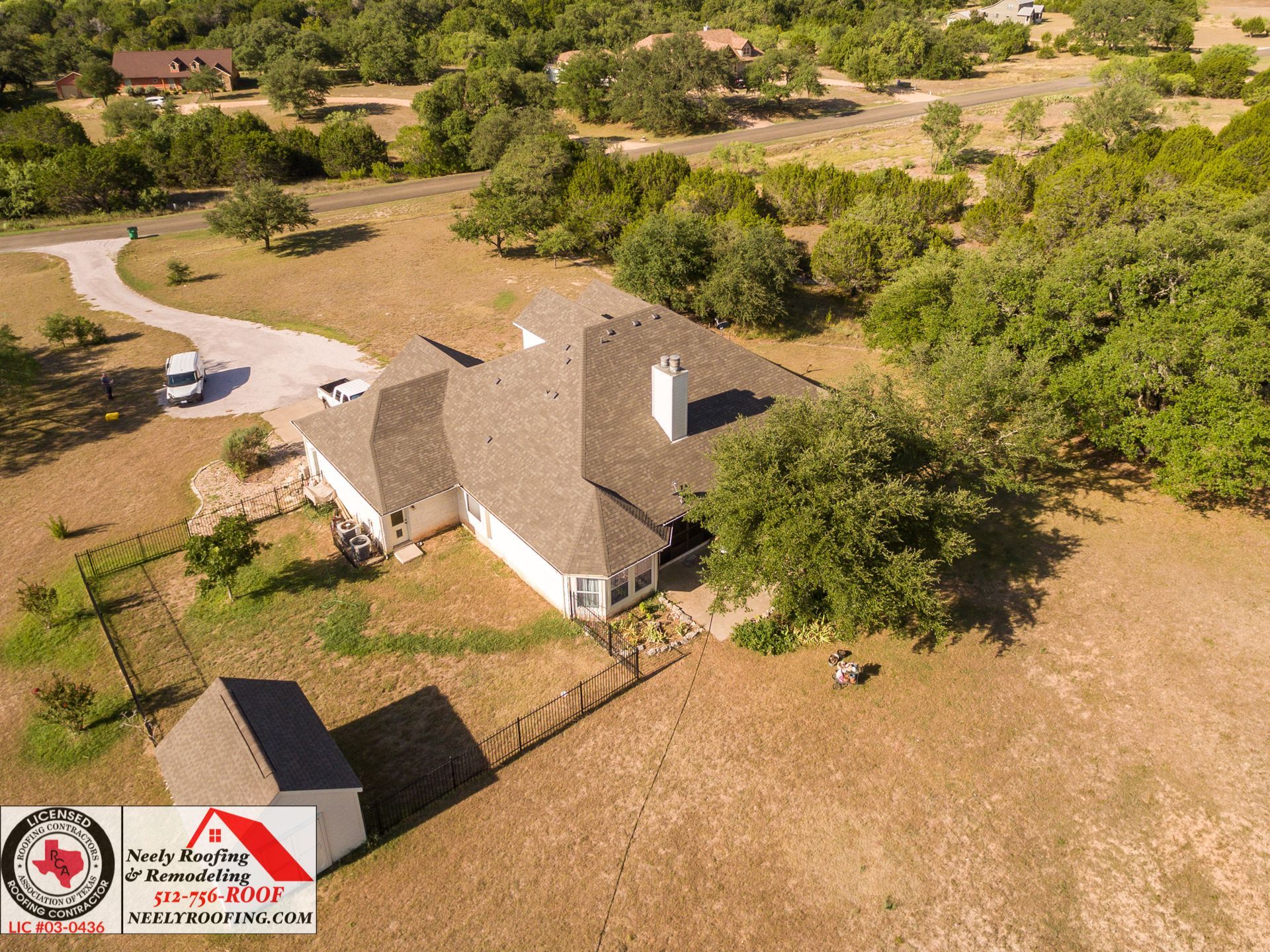 Aerial view of a tan-roofed house with a driveway and surrounding trees in a dry, rural landscape