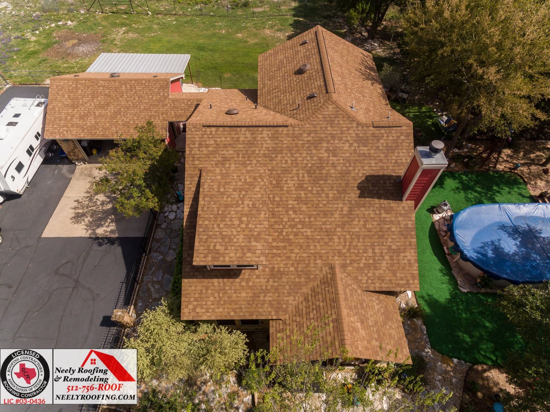 Aerial view of a brown-shingled house with a driveway and a blue pool beside trees.