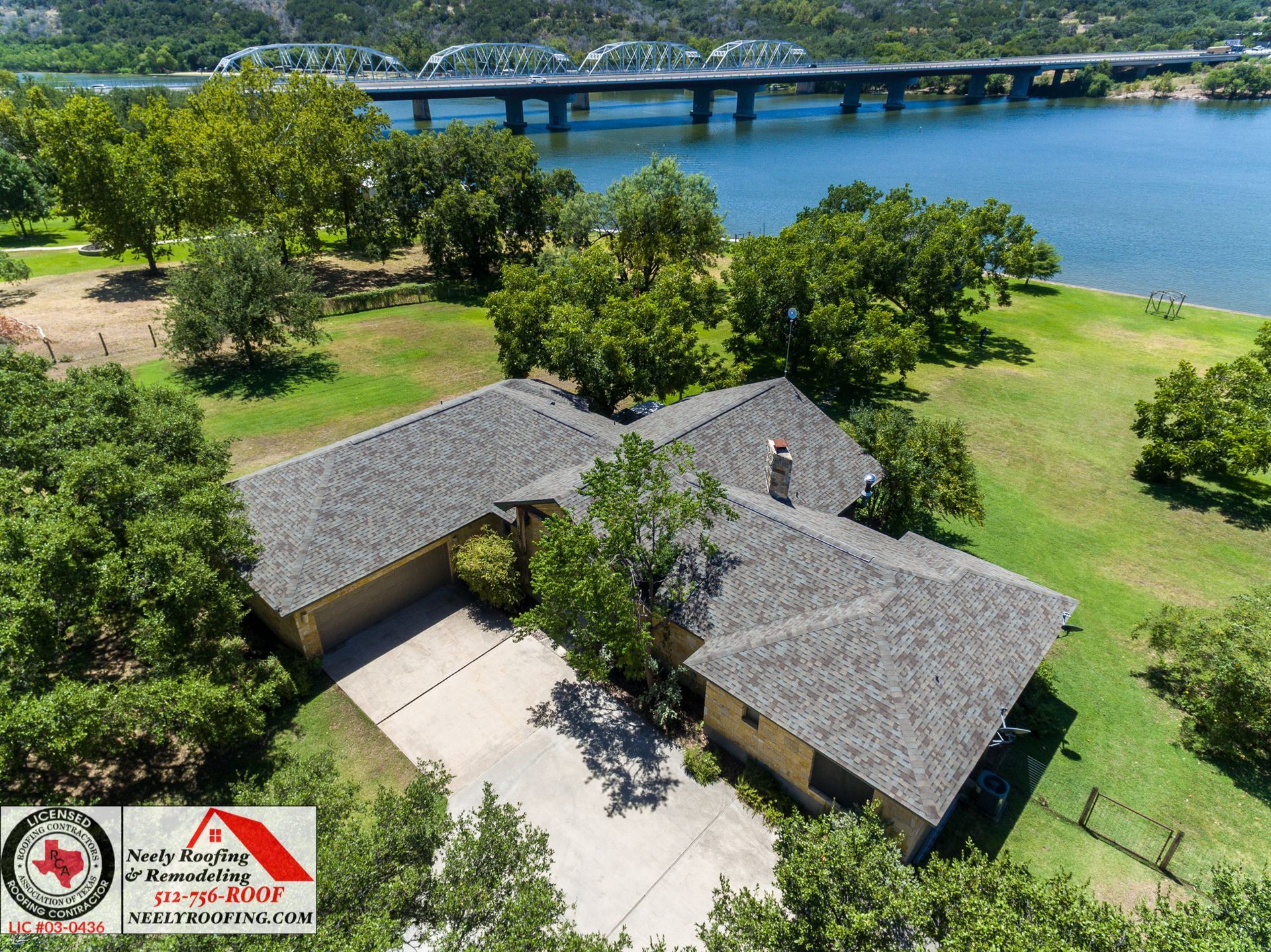 Aerial view of a house with gray roofs beside a green lawn and blue water, with a bridge in the background