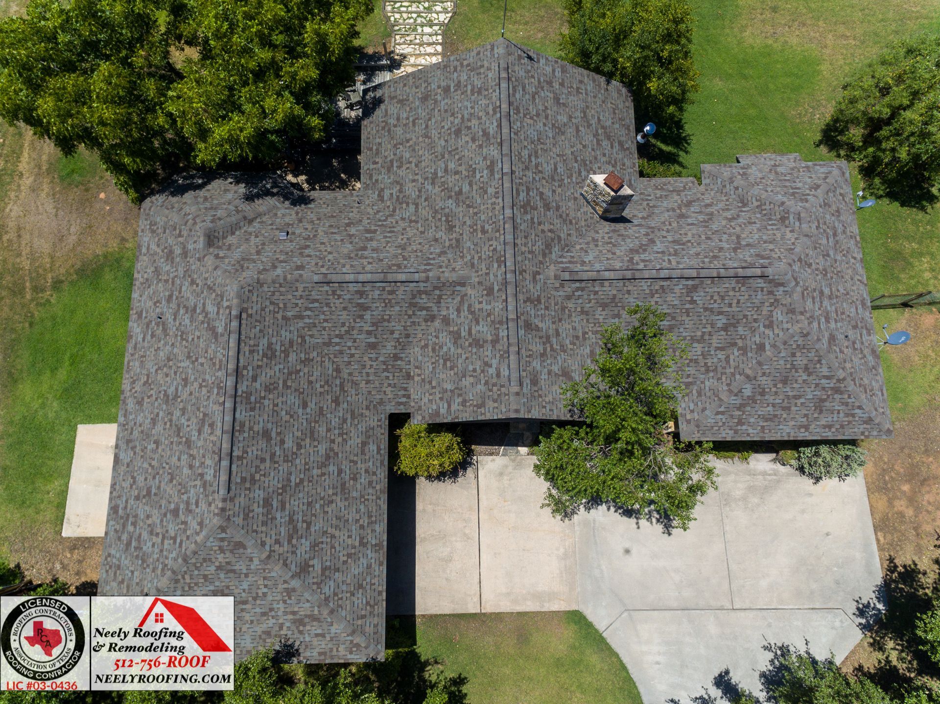 Aerial view of a house with a gray shingled roof and driveway, surrounded by green lawn and trees.