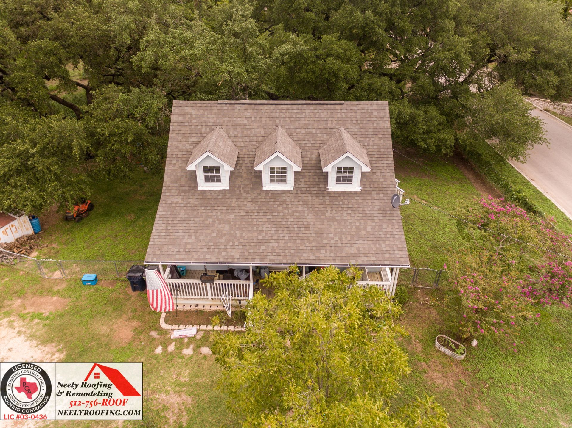 Aerial view of a gray-roofed house with dormer windows, surrounded by trees and grass.