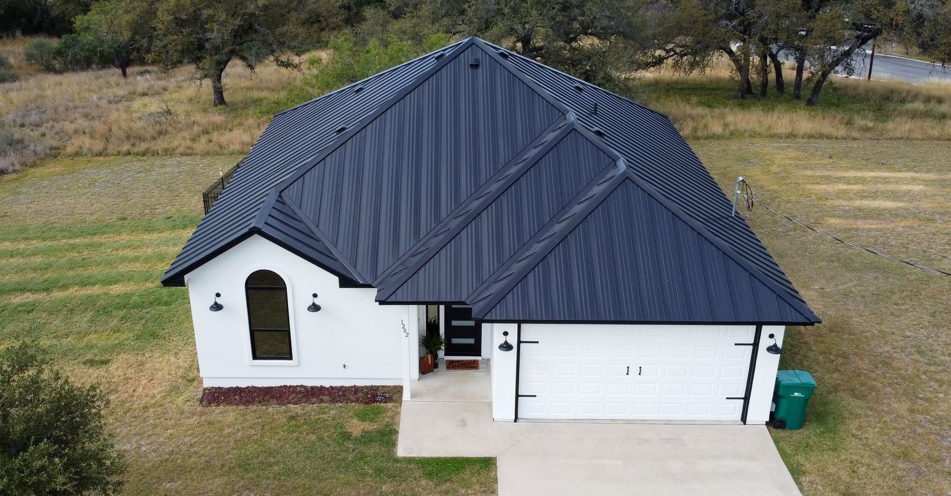 Aerial view of a white house with a dark roof, garage, and driveway in a grassy rural setting.