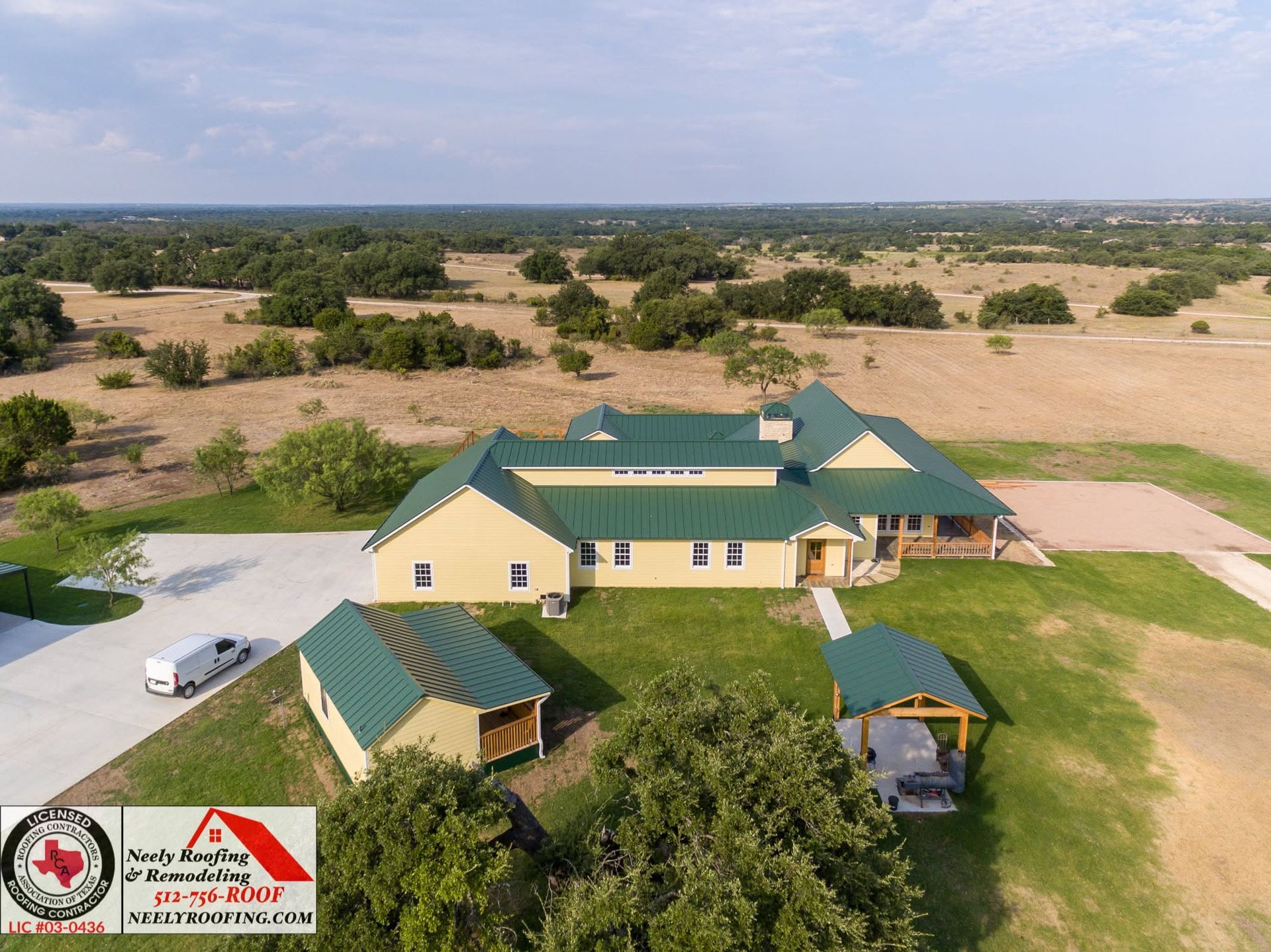 Aerial view of a yellow house with green roofs, surrounded by trees and open dry fields.