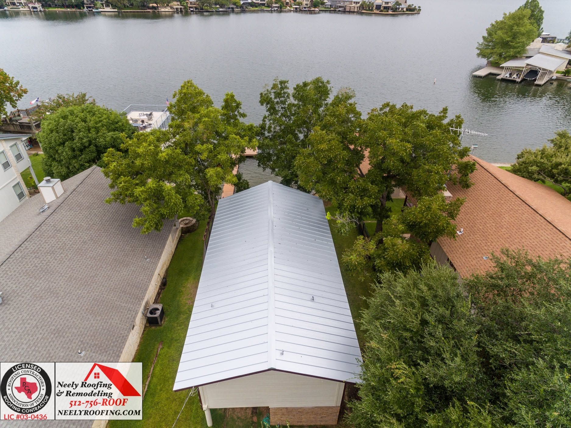 Aerial view of a lakeside property with trees, rooftops, and dock along the shore