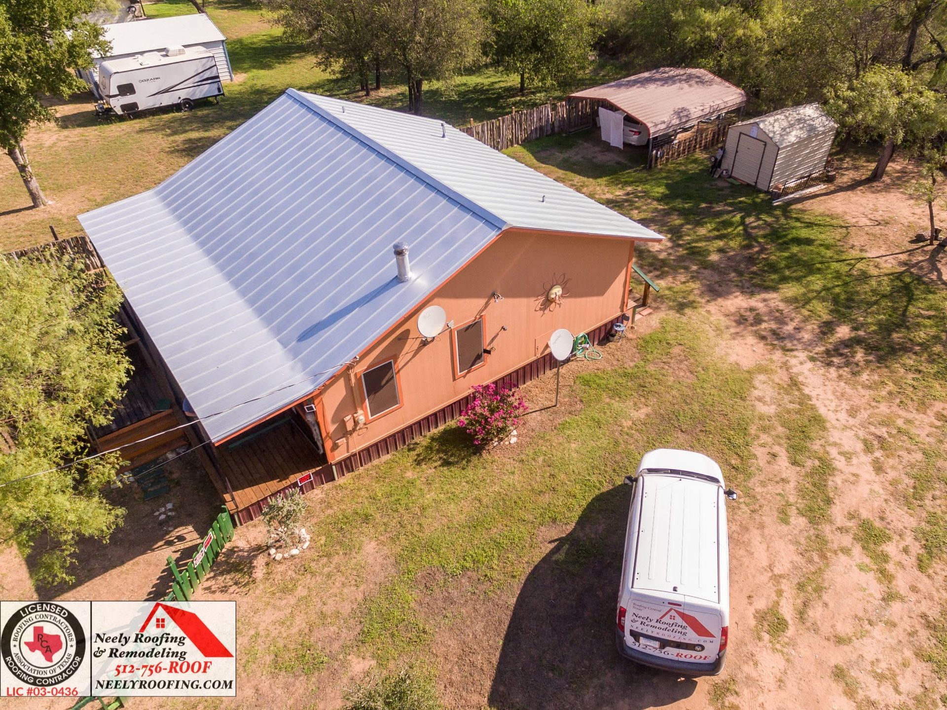 Aerial view of a brown house with a metal roof, surrounded by trees and parked vehicles.