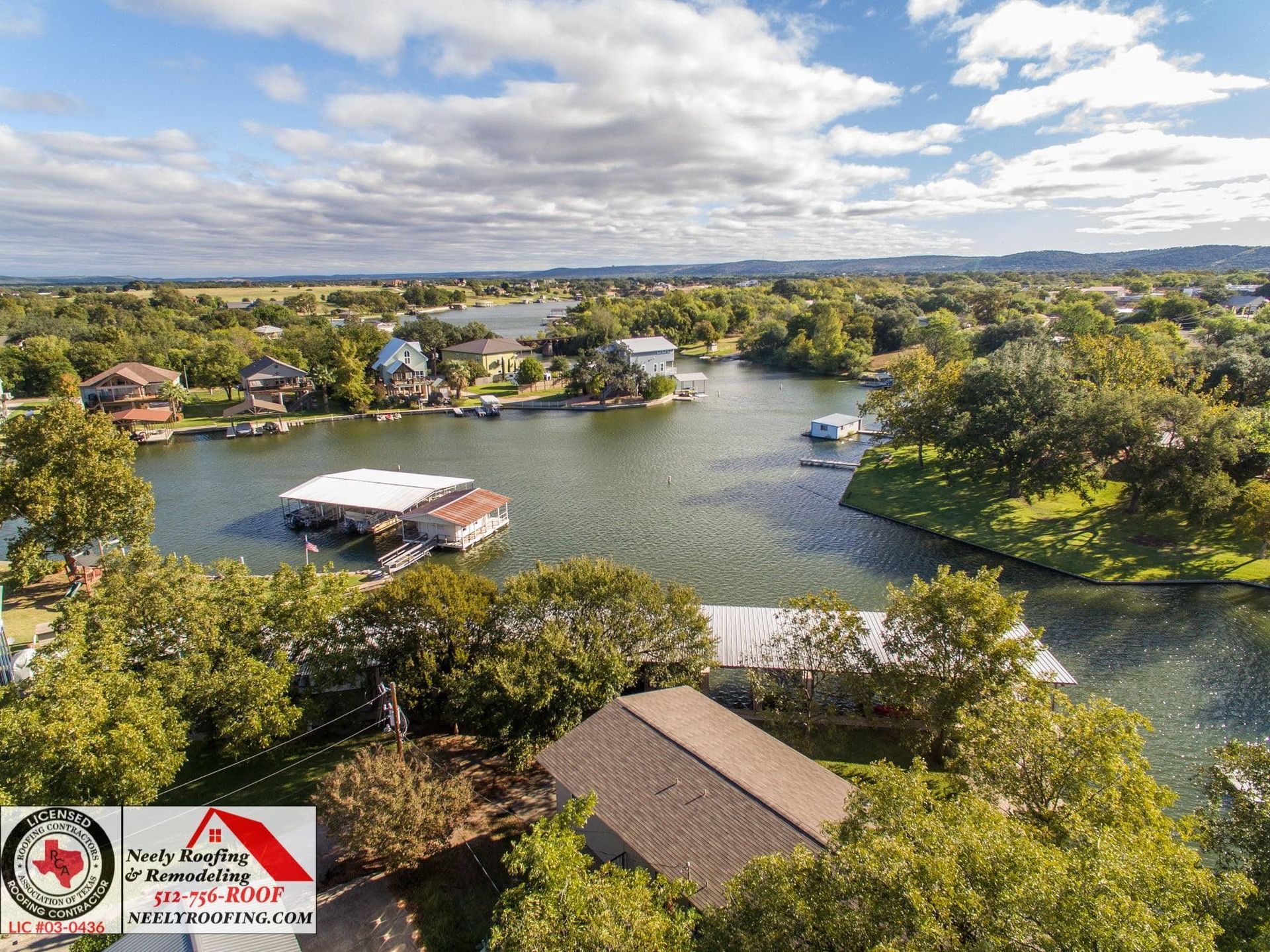 Aerial view of a lakeside village with houses, docks, and trees under a partly cloudy sky.
