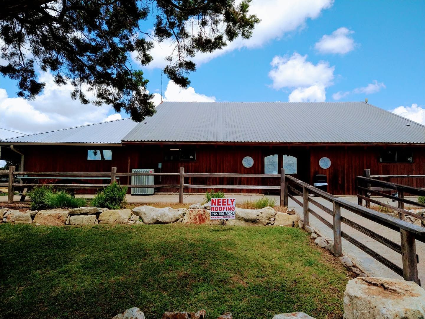 Red barn-like building with a metal roof, wooden porch, and fenced grassy yard under a blue sky.