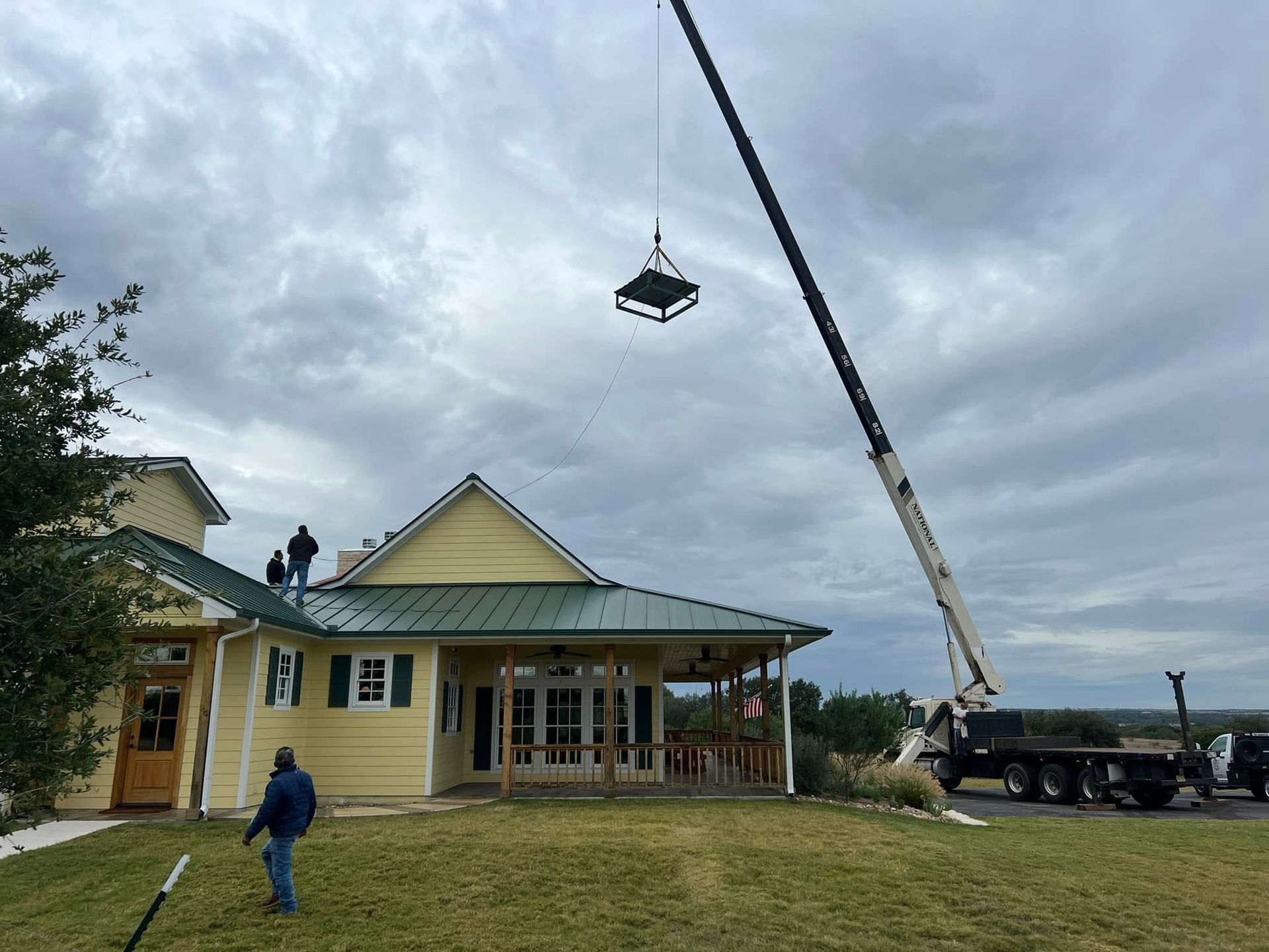 Crane lifting a roof truss over a yellow house under construction, with workers nearby.